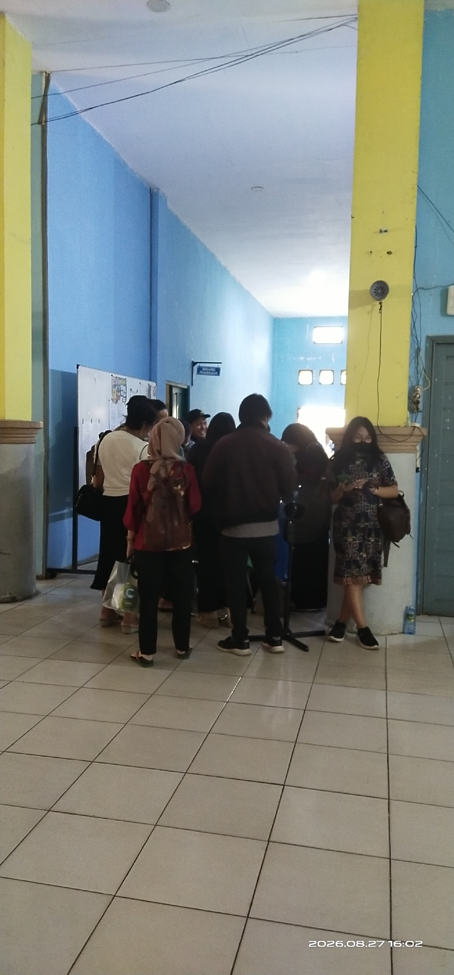 In a well-lit indoor hallway with blue and yellow painted walls, a group of approximately ten people are gathered. The floor is tiled with a light-colored, square pattern. Several individuals are standing and conversing, with some holding bags or backpacks. One person in the foreground is wearing a patterned dress and holding a phone. Another person, wearing a red shirt and headscarf, has a backpack. A sign is visible on the blue wall with text that appears to be in Indonesian. The overall scene suggests a casual gathering or waiting area. The timestamp "2026.08.27 16:02" is present in the lower right corner of the image. While the image is located in the city of Tamiang Layang, Indonesia, specific details about the exact location beyond it being an indoor hallway are not discernible from the visual information provided.