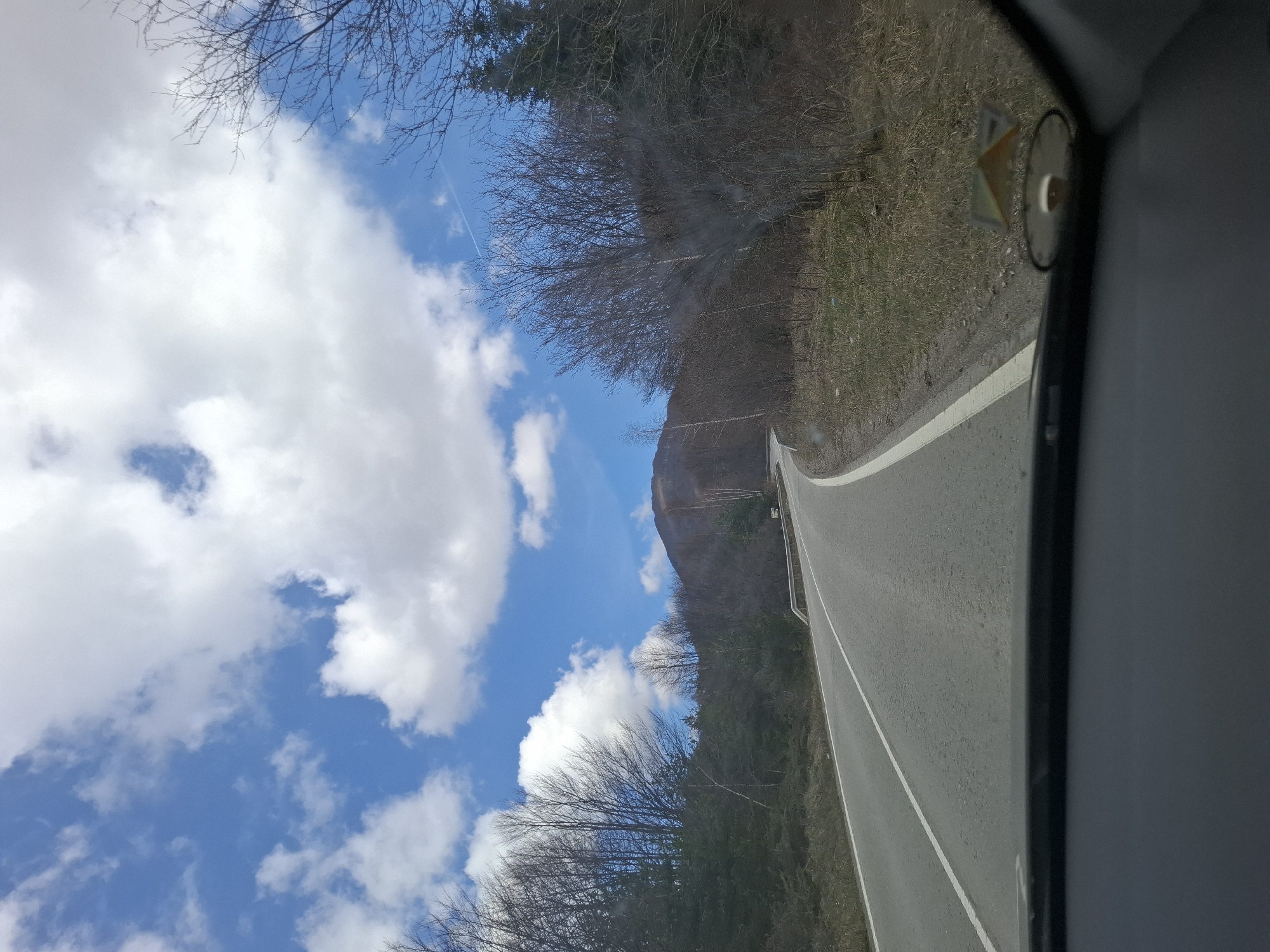 The image depicts a view from inside a vehicle, likely a car, driving on a road in what appears to be a mountainous area near Gintsi, Bulgaria. The road is paved with a white line marking the lanes. To the side of the road, there's sparse vegetation and bare trees, suggesting either late autumn or early spring. In the distance, a mountain is visible under a sky with blue patches and scattered white clouds.