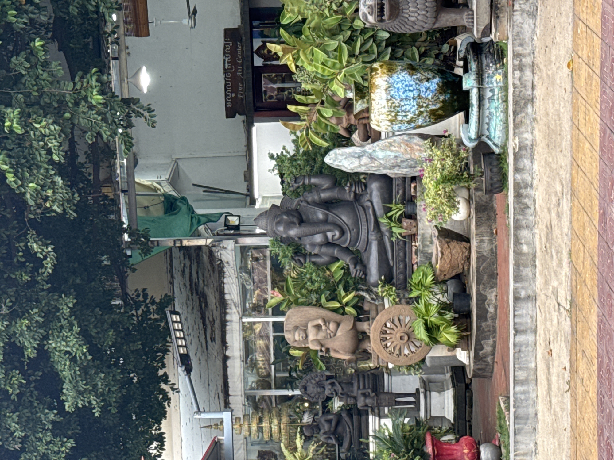The image depicts an outdoor display of statues and decorative objects in front of a building, likely a shop or gallery, located in Khan Daun Penh, Cambodia. A large, dark stone statue of Ganesha, the Hindu deity with an elephant head, is prominently positioned in the center, facing the viewer. To the left of Ganesha is a statue of a figure riding a bird, resting on a large stone wheel. Several other dark stone statues of human figures in seated and standing poses are arranged behind these central pieces, some appearing to be monks or deities. The display is surrounded by lush green foliage and potted plants.
In the background, the building has large glass windows, suggesting it's a commercial establishment. A sign above the entrance, partially obscured by the foliage, appears to be in Khmer script and also reads "Fine Art Center" in English. A light fixture hangs from the ceiling above the entrance. To the right of the display, a decorative fountain with a blue and green glaze is visible, and further to the right, a stone sculpture of a lion's head is placed. The ground in the foreground is paved with yellow and red tiles, with a concrete ledge separating it from the display area. The overall scene suggests a cultural or artistic retail environment.