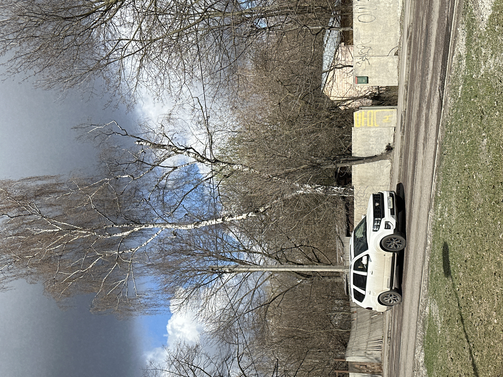 The image shows a white SUV parked on a street beside a sidewalk with patches of grass, near a building. Trees with bare branches are visible, suggesting late autumn or early spring. The sky is partly cloudy with dark, ominous clouds looming. The license plate on the vehicle is visible. Graffiti is visible on the concrete blocks along the sidewalk. The location is in the city of Moscow, Russia based on the vehicle license plate.