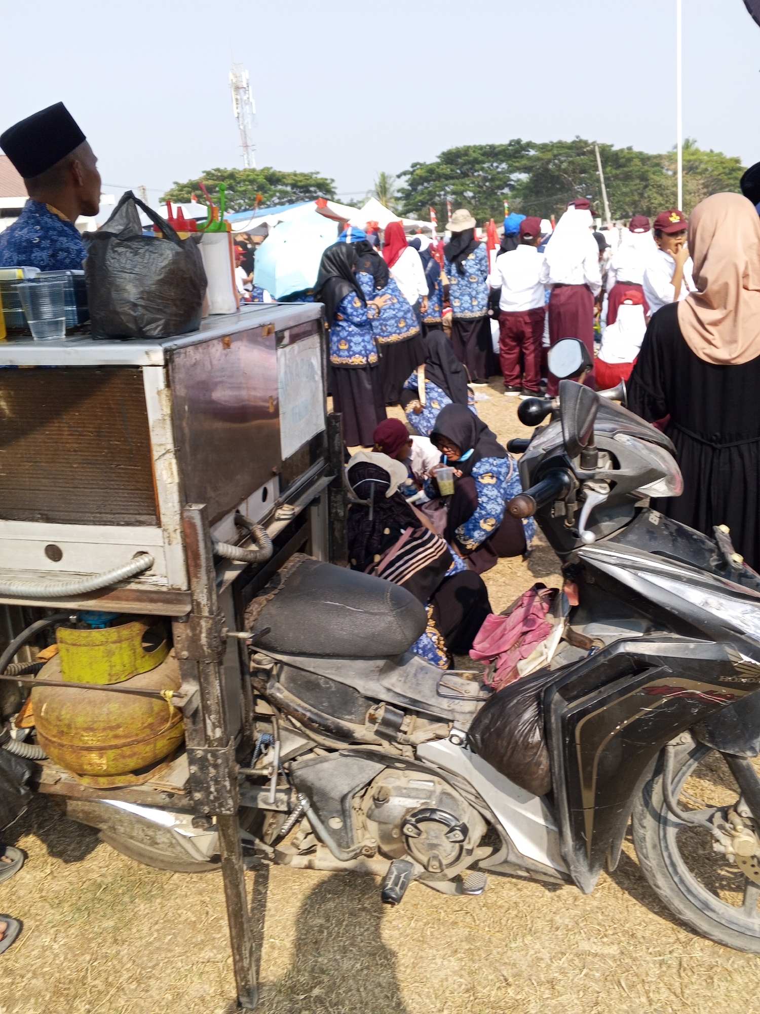 The image depicts an outdoor scene in Ranjeng, Indonesia, likely during an event or market. In the foreground, a modified motorcycle serves as a mobile cart. The motorcycle is black and white, with a worn black seat and various attachments. Attached to the rear of the motorcycle is a metallic cart, which appears to be a makeshift refrigerator or cooler, fitted with a ventilation grill on its left side and some pipes. A yellow gas cylinder is visible beneath the cart.

Numerous people are present in the background, many of whom are wearing uniforms. Some are dressed in blue and white uniforms with intricate patterns, while others wear maroon and white attire, including maroon skirts and white shirts or maroon caps. Several individuals are wearing headscarves, with one woman in a light brown hijab prominent on the right side of the frame. The people are gathered, some standing and others seated on the ground, suggesting a public gathering or a commercial setup. The ground is covered with dry grass, and in the distance, trees and a cellular tower are visible under a clear sky. The overall atmosphere suggests a community event or a busy marketplace.