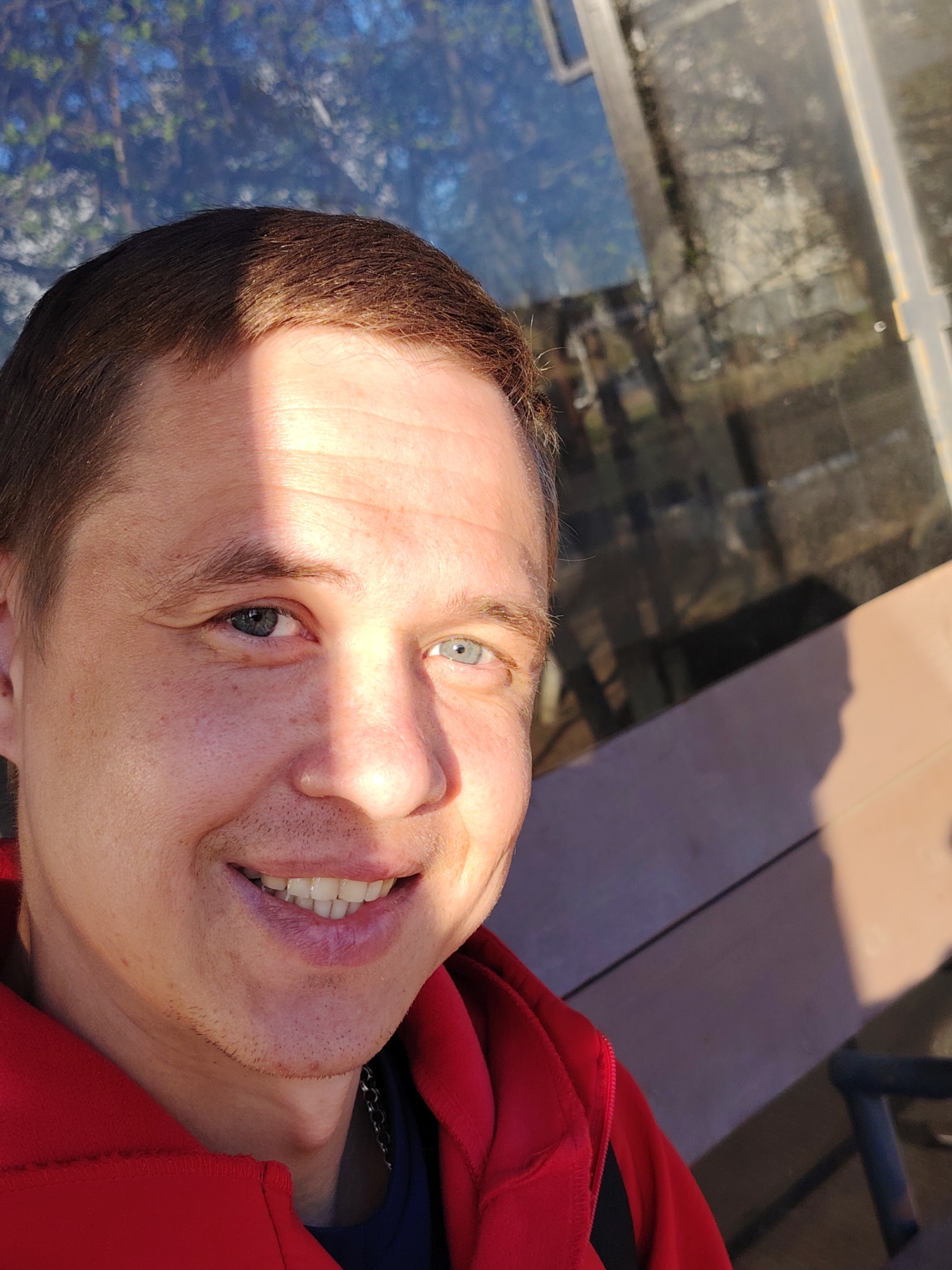 A selfie depicts a man with short brown hair and blue eyes smiling at the camera. He is wearing a red jacket over a dark blue shirt and a silver chain necklace is visible. A vertical strip of bright sunlight falls across his forehead and nose. Behind him is a window reflecting trees and sky. The location is Chaikovsky, Russia.
