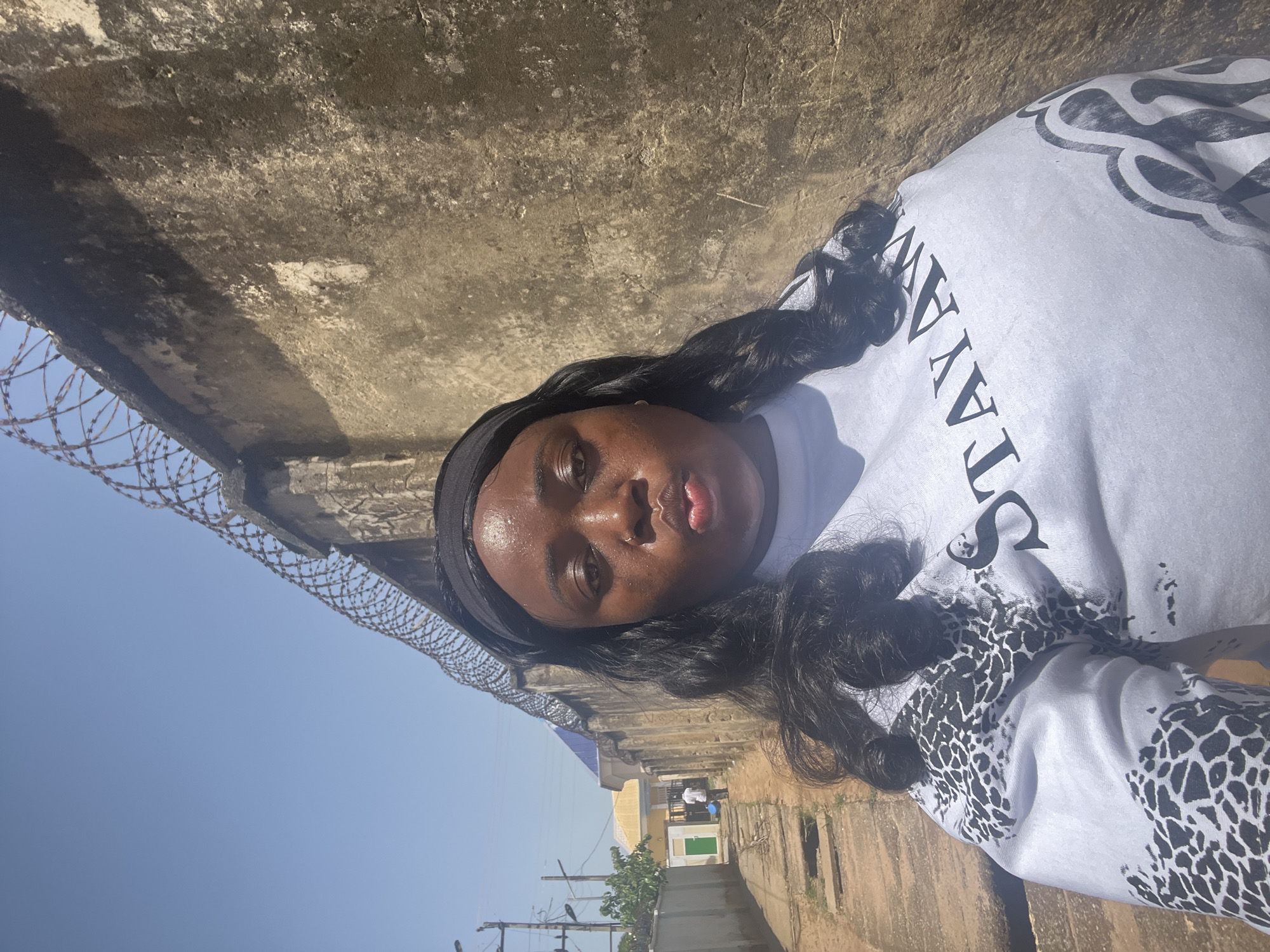 A medium shot of a Black woman with long, wavy black hair, wearing a black headband and a white sweatshirt with black text and an animal print. She is standing in an alleyway next to a tall, concrete wall topped with razor wire. The sky is light blue. In the background, a building with a yellow facade and a green door is visible, along with two people and utility poles. Based on the buildings, this alley appears to be located within the city of Abuja, Nigeria.