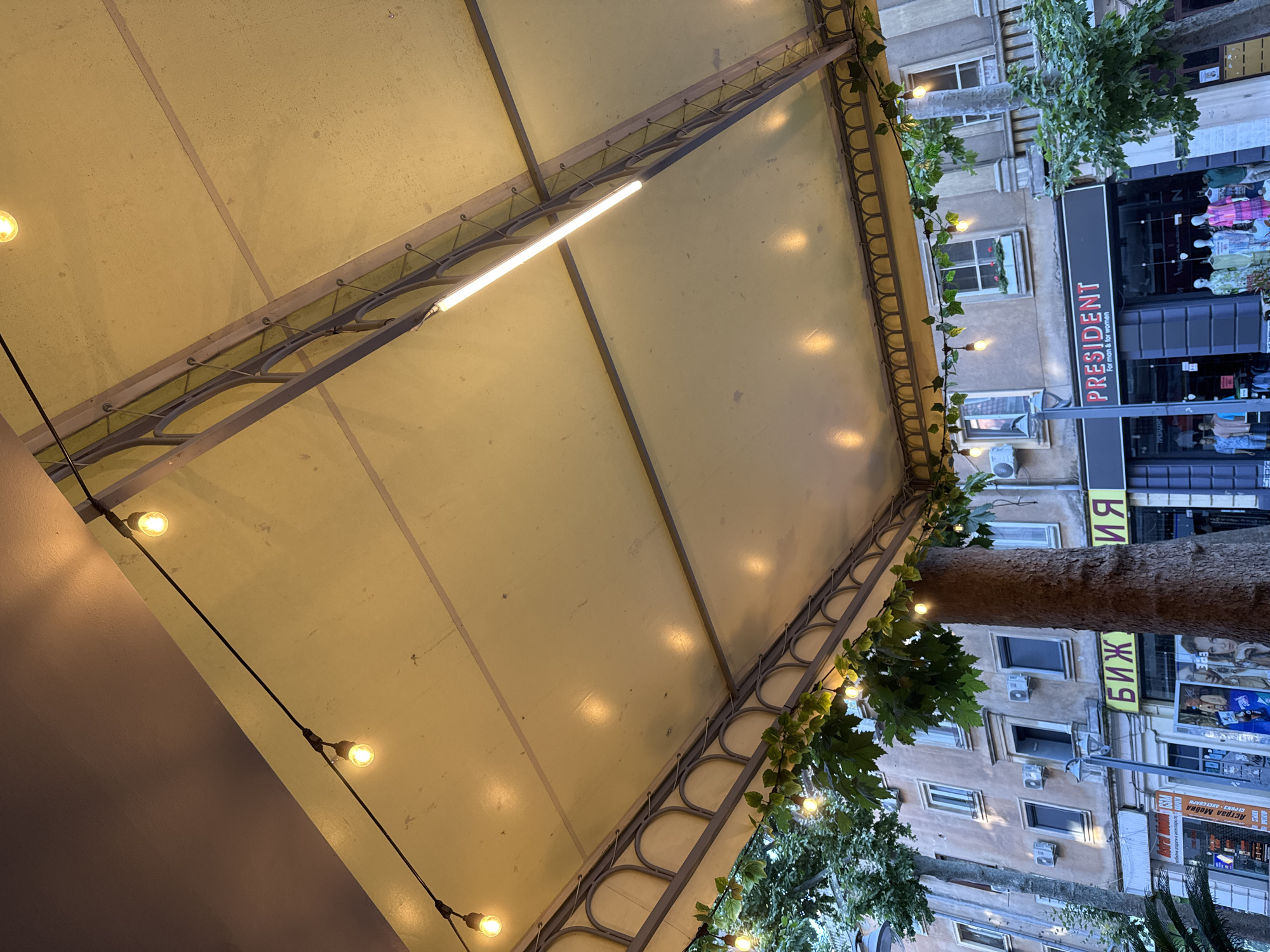 The image shows an outdoor cafe or restaurant with a light-colored awning illuminated by string lights and a fluorescent tube light. Greenery with small lights is draped along the edge of the awning. Below the awning, there are buildings with shops. One shop's signage reads "PRESIDENT For man & for women." Other visible signs include Cyrillic script, one of which reads "БИЖУТЕРИЯ," which translates to "Jewelry." Several trees are visible, including a large one supporting the awning. The architecture suggests a European city, and the Cyrillic script indicates a location in a country using that alphabet. Based on previous knowledge, this scene is located in Burgas, Bulgaria. The lighting and time of day suggest it might be evening or a cloudy day. No people are clearly visible in the foreground.