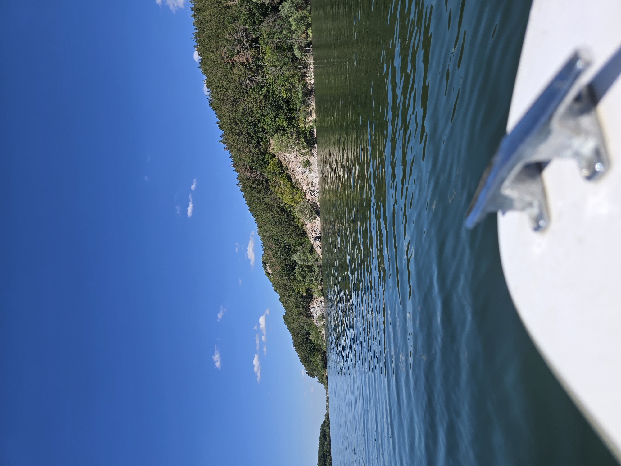The image depicts a serene lake scene under a clear blue sky with scattered white clouds. The foreground features the edge of a white boat, with a silver cleat prominently visible and out of focus. The middle ground is dominated by the dark, rippling surface of the lake, reflecting the surrounding landscape. The background showcases a lush, tree-covered hillside that slopes down to the water's edge. Rocky outcrops are visible on the hillside, and a small dark opening, possibly a cave or tunnel, is present on the right side of the hill near the water. The overall impression is one of natural tranquility and a recreational outing on the water.