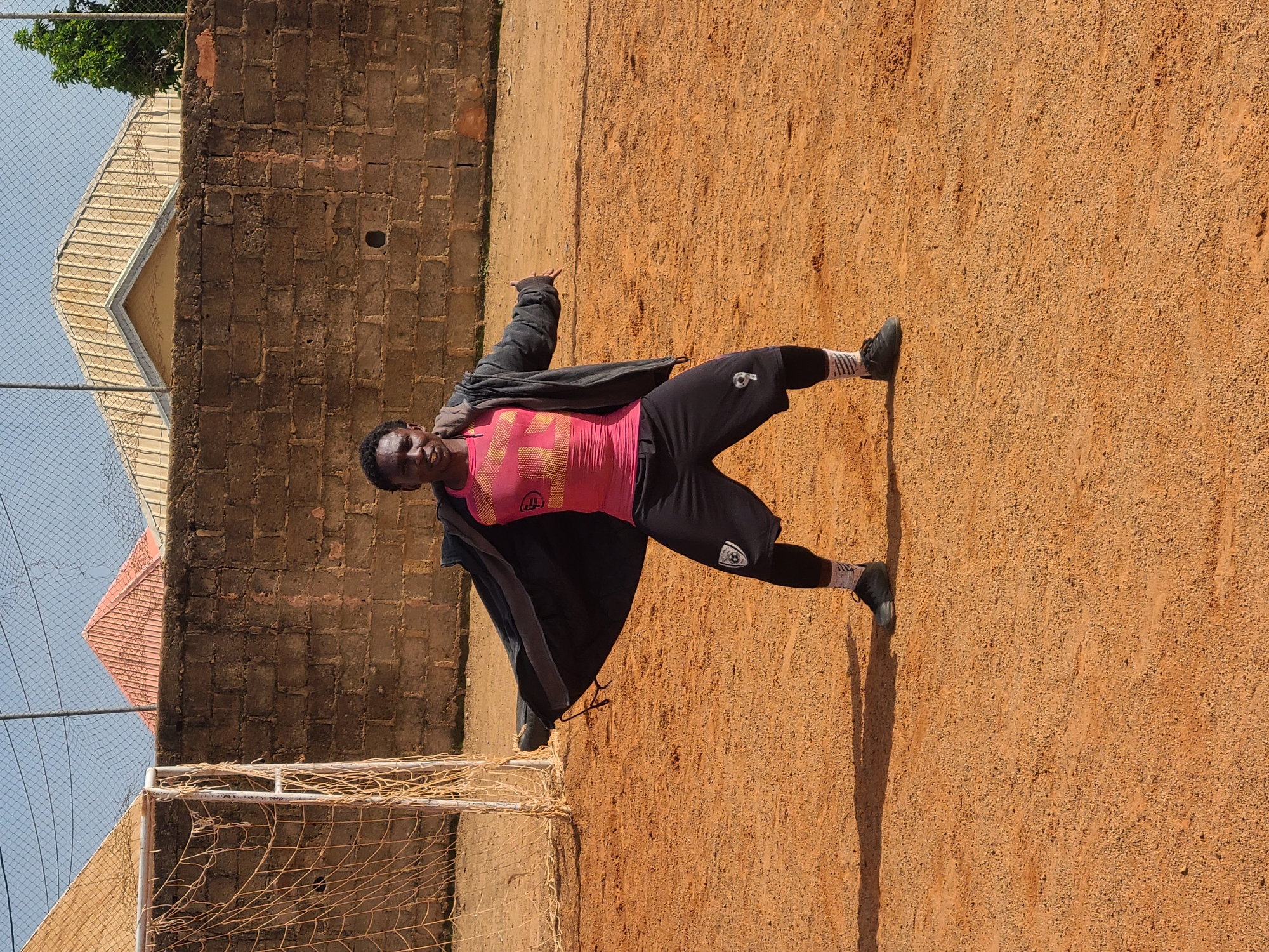 A young Black man stands on a dirt field in Barakin Lerawi, Nigeria, with his arms outstretched. He is wearing a pink t-shirt with a yellow geometric pattern, black athletic shorts over black leggings, and black sneakers. His dark jacket is draped open over his shoulders. Behind him, a brick wall runs horizontally across the frame, topped by a chain-link fence. Beyond the fence, several buildings with peaked roofs are visible, as is a segment of a soccer goal with a net. The ground is an even, reddish-brown dirt. The lighting suggests it is daytime.