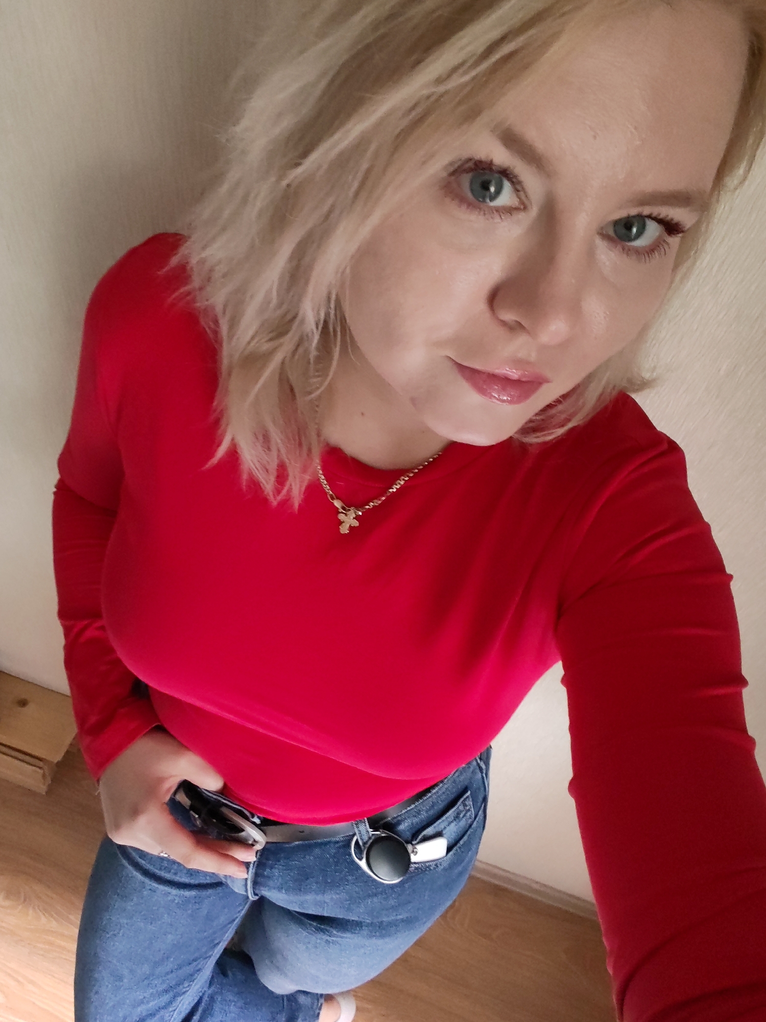 A young woman with blonde hair and blue eyes is taking a selfie. She is wearing a red long-sleeved shirt and blue jeans. She has a silver necklace with a cross pendant and a belt with a silver buckle. She is holding what appears to be a smartphone in her right hand, and her left hand is resting on her hip. The background appears to be an indoor setting with a light-colored wall and wooden floor. There is no discernible information in the image to suggest a location in Izhevsk, Russia.