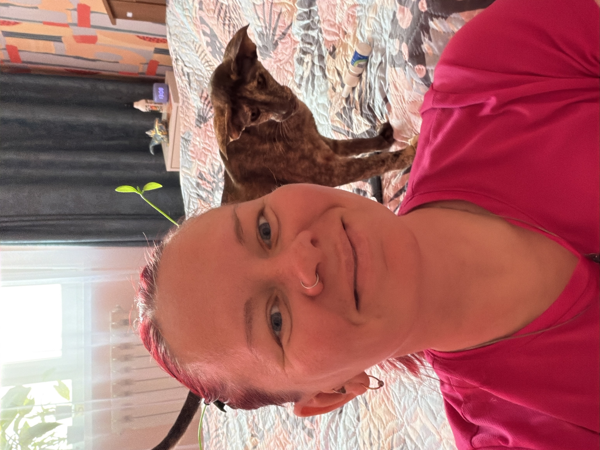 The image shows a person, likely female, with pinkish-red hair, blue eyes, and a nose ring, wearing a bright pink shirt. She is indoors, lying on a bed with a patterned comforter. Behind her, a brown tabby cat is positioned on the same bed. There's a window with blinds in the background, revealing some greenery outside. Next to the bed, there's a small table with an object on it. Based on the information provided, the location is Chaikovsky, Russia.