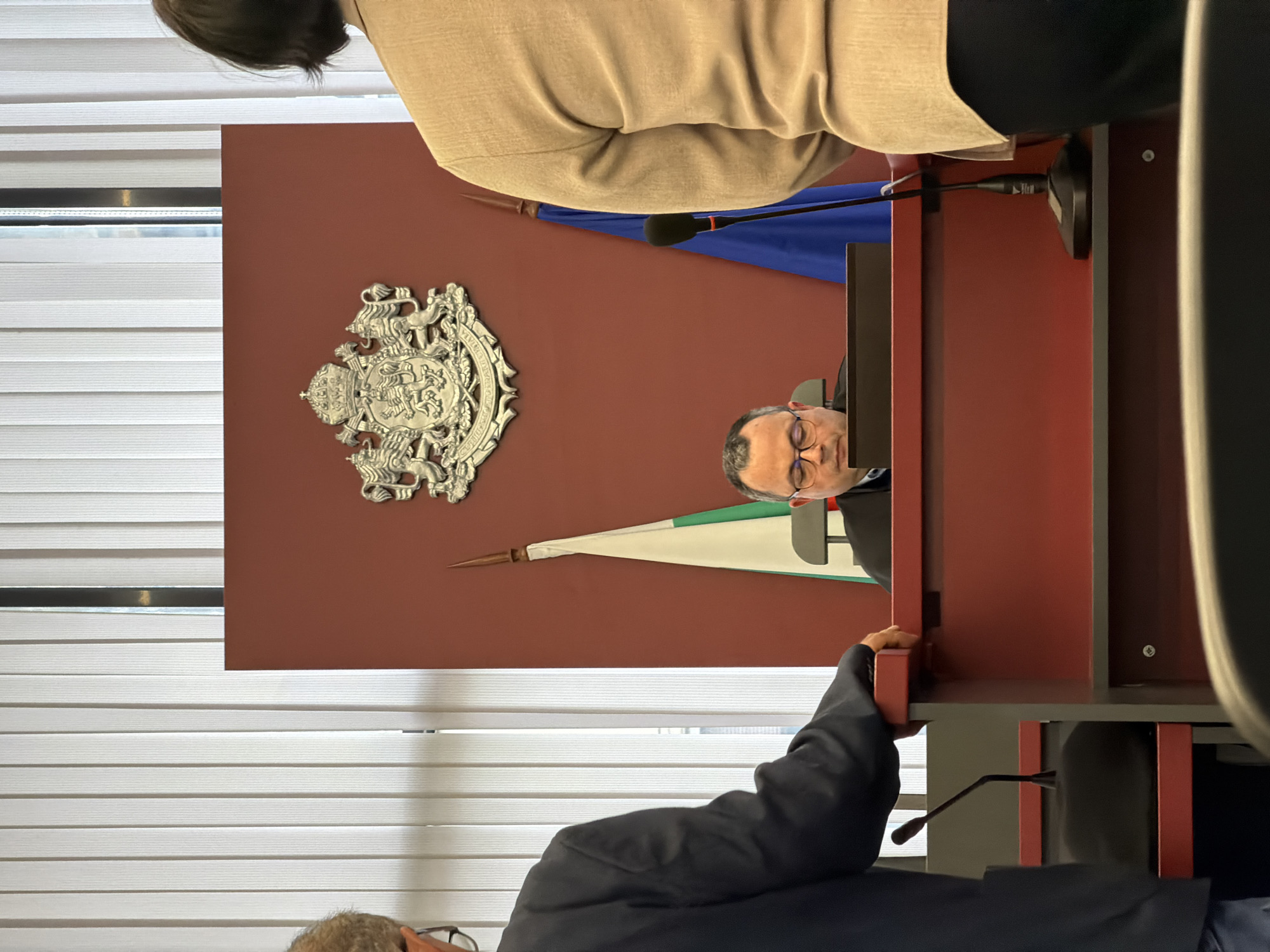A medium shot captures a man with glasses looking into a monitor from behind a red lectern. The wall behind him is also red and features a silver emblem on the upper-left and a flag pointing toward the left center. A dark-haired woman in a tan blazer and a gray-haired man in a dark blazer are partially visible in the upper left and lower center, respectively. White blinds are visible on the left side of the frame. The location is in Plovdiv, Bulgaria, based on the flag of Plovdiv in the background.