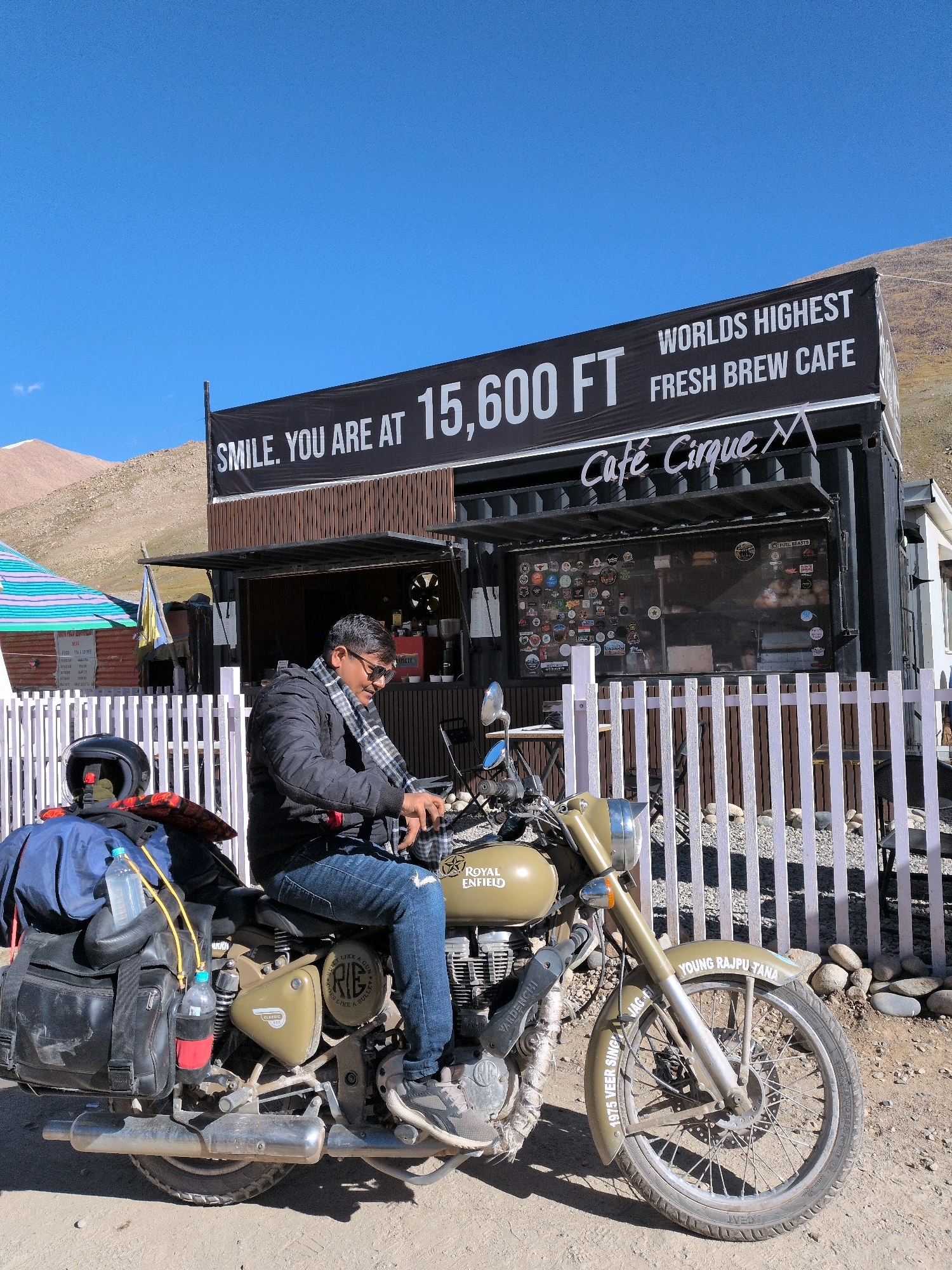 A man sits on a Royal Enfield motorcycle parked in front of a cafe at Khardung La, India, marked as the "World's Highest Fresh Brew Cafe" at 15,600 feet. The man, wearing a black jacket, blue jeans, sunglasses, and a scarf, is looking down at the motorcycle's controls. The motorcycle is loaded with luggage, including saddlebags, a backpack, and water bottles. Behind the motorcycle, a white picket fence separates the cafe from the road. The cafe itself is a shipping container converted into a business, with a banner across the top and a window displaying various stickers. The background shows a mountainous landscape under a clear blue sky.