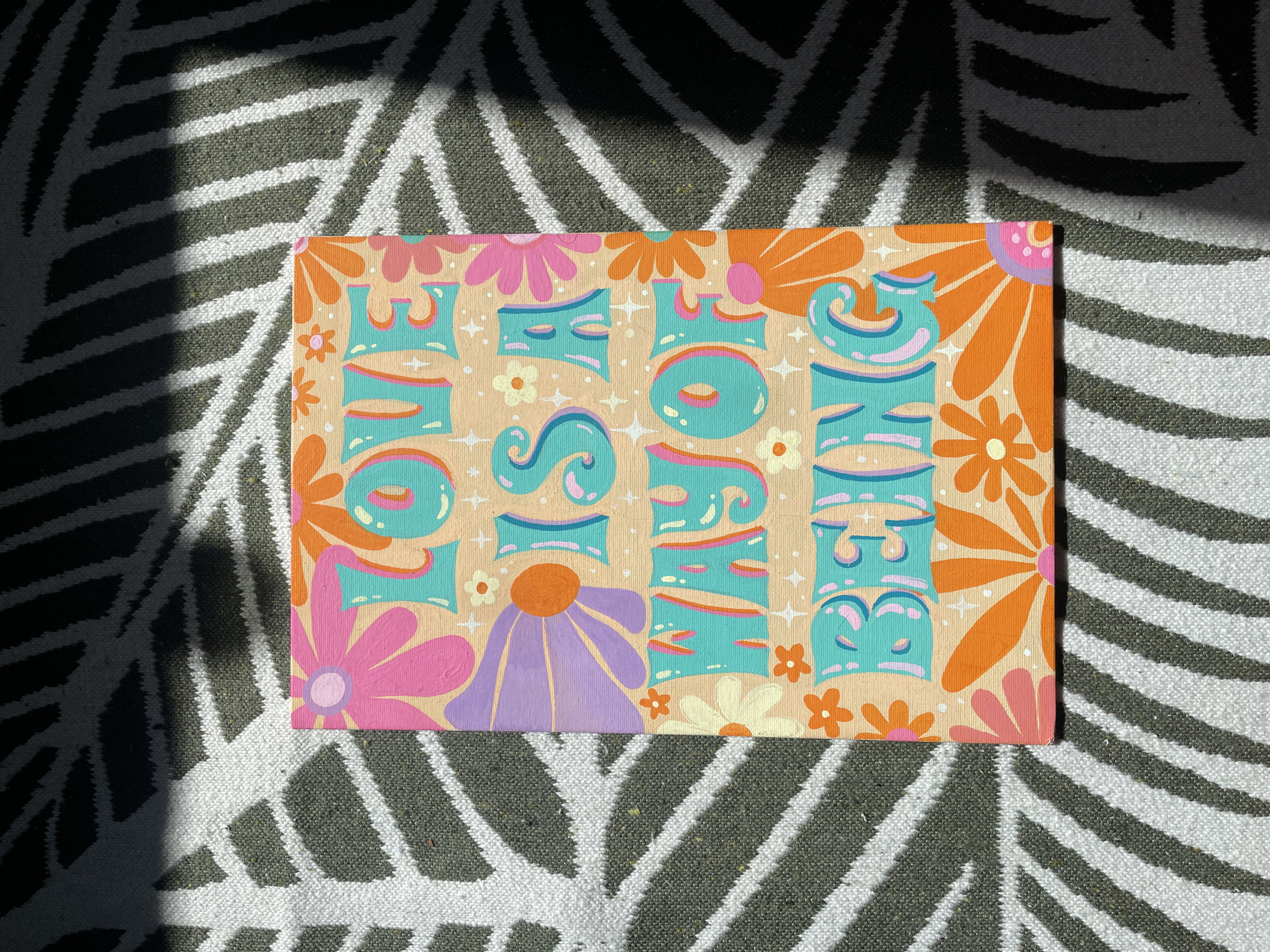 The image shows a painting with the words "LOVE IS A WAY OF BEING" written in colorful, retro-style lettering. The painting is adorned with stylized flowers in shades of pink, orange, purple, and cream. The background of the painting is a light peach color. The painting is resting on a rug with a black and white striped pattern. Sunlight is casting shadows across the rug and the painting, creating a dappled effect. The exact location in the city of Kopeysk, Russia, is not discernible from the image.