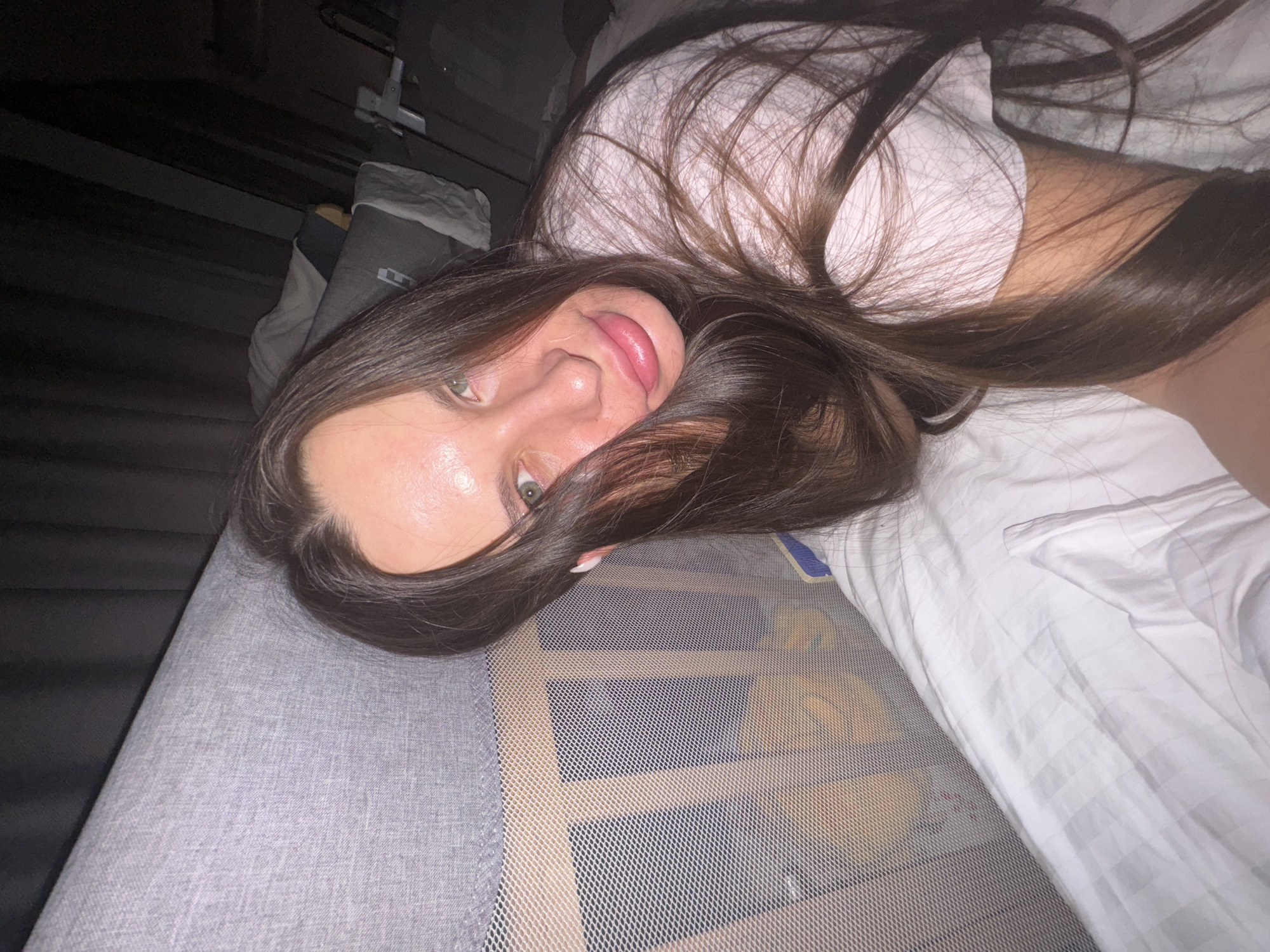 The image features a young person, likely female, with long, straight dark brown hair and greenish eyes, lying on a bed and looking directly at the camera. She has a slight, relaxed smile. She is wearing a white t-shirt.
The setting is an indoor bedroom, characterized by soft, possibly dim, lighting. The bed is covered with white sheets that display a subtle woven or striped pattern. To the person's left, a grey upholstered surface, likely a headboard or side of the bed, is visible. To her right, a light-colored, fine-mesh barrier, possibly a bed rail or part of a cot, extends across the mid-ground.
A notable object in the upper left background is a dark-colored item resembling an assault rifle, which appears to be mounted or leaning against a dark wall or piece of furniture. No visible text is discernible on the person's clothing or the immediate bed linens. The overall impression is one of a casual, private moment.