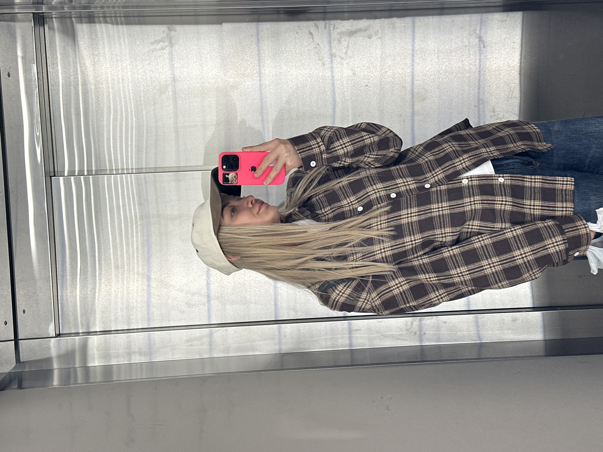 A woman is taking a selfie in an elevator. She is wearing a plaid shirt, jeans, and a baseball cap. She is holding a pink iPhone. The elevator is made of stainless steel. The location in the city of Budva, Montenegro, cannot be determined from the image.