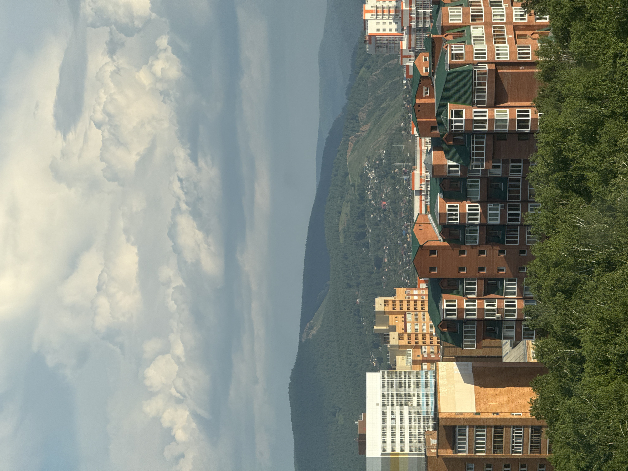 The image depicts a cityscape of Krasnoyarsk, Russia, featuring a cluster of modern apartment buildings in the foreground, characterized by brick facades and green roofs. Behind these structures, a verdant hillside rises, dotted with smaller houses and trees. In the distance, larger, forested mountains form the backdrop, under a sky filled with dramatic, illuminated clouds. The scene is captured during daylight, with shadows suggesting it is late afternoon or early evening. No people are visible in the image.