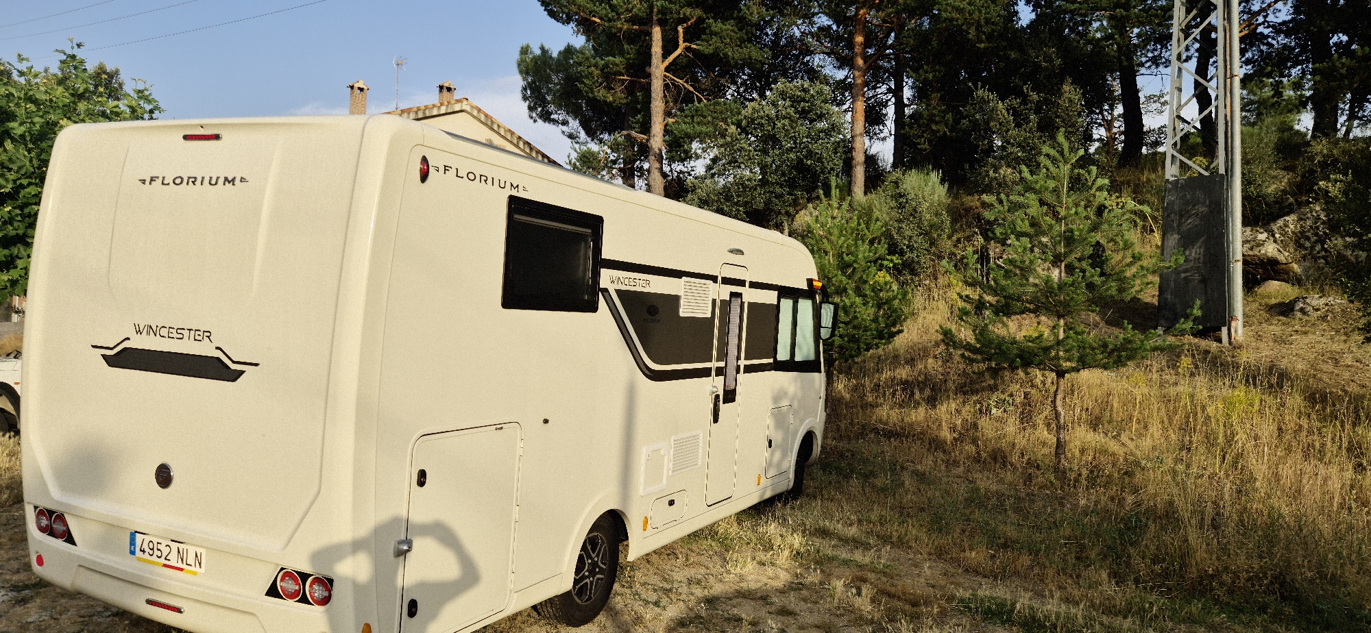 A cream-colored motorhome is parked on a dirt road next to dry grass and trees under a clear sky. The motorhome has the brand name "FLORIUM" and "WINCHESTER" visible on its exterior. The license plate of the motorhome reads "4952 NLN". The motorhome is parked on a slight incline, with a metallic pole and some rocks visible in the background. The scene appears to be in a rural or natural setting, with no people visible in the image.