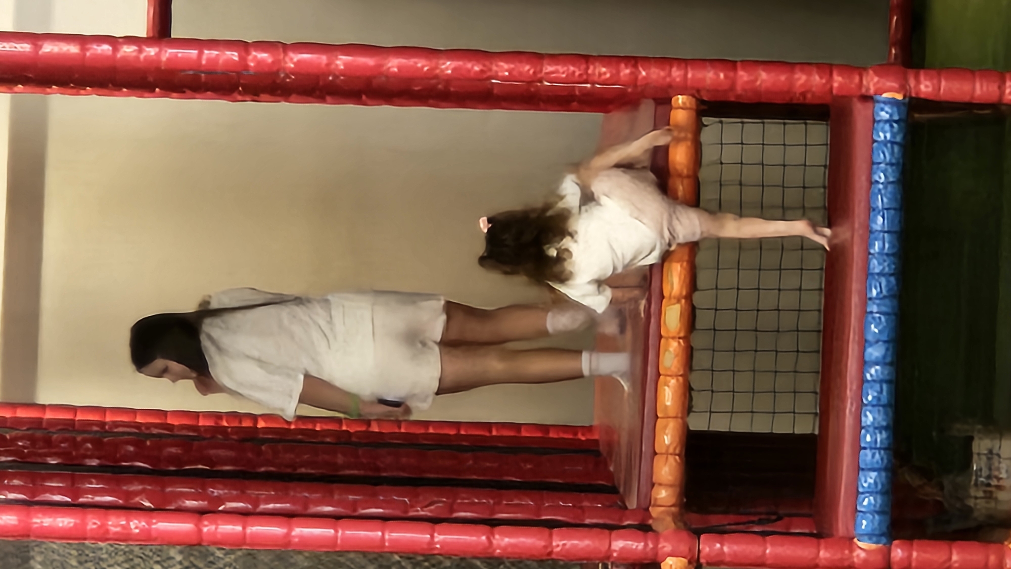 In an indoor play area, two individuals are visible on a multi-level soft play structure. A taller individual, likely a young woman, stands facing away from the camera, wearing a white t-shirt and white shorts. Her right leg is bent at the knee, and she appears to be looking towards the left. Beside her, a younger child is positioned lower on the structure, facing away from the camera and to the right. The child is wearing a white t-shirt and light-colored shorts and is in the process of climbing or moving. The play structure features red padded vertical poles and orange and blue padded horizontal surfaces, with a black net visible in the background. The overall environment is consistent with a children's entertainment center. The location is not specified to be in Bratislava, Slovakia.
