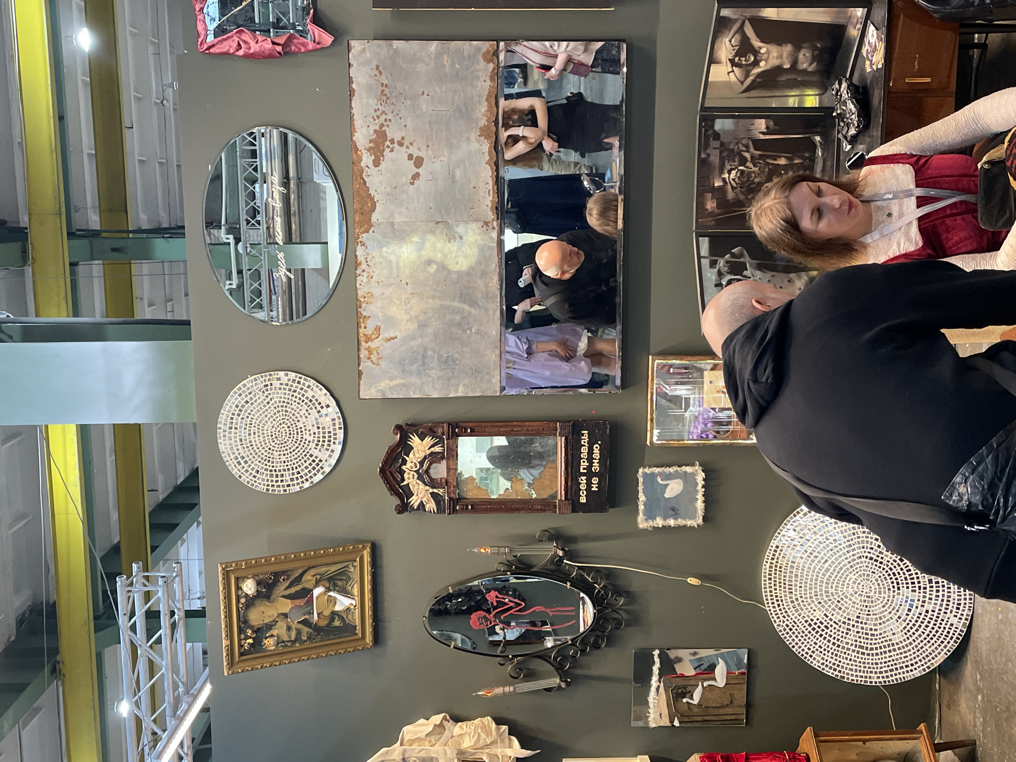 The image shows a section of a wall in what appears to be an exhibition or gallery space. The wall, painted in a muted grey-green, is adorned with a diverse collection of decorative mirrors and artworks. Several mirrors of varying shapes and sizes are present, including oval, rectangular, and square mirrors, some with ornate frames. One mirror has text in Russian. There's also a framed painting and decorative wall art, including a circular piece made of small mosaic tiles. The ceiling of the space is visible in the top left corner, featuring industrial-style metal beams and fluorescent lighting.
Two people are in the frame. One woman is wearing a red dress with white trim. A man in a black hooded sweatshirt is positioned facing away from the camera. They appear to be at the show or gallery.
Based on the objects and the presence of Russian text, the location is likely in Saint Petersburg, Russia. The content on the wall suggests a display of art or decorative pieces, potentially as part of an exhibition.