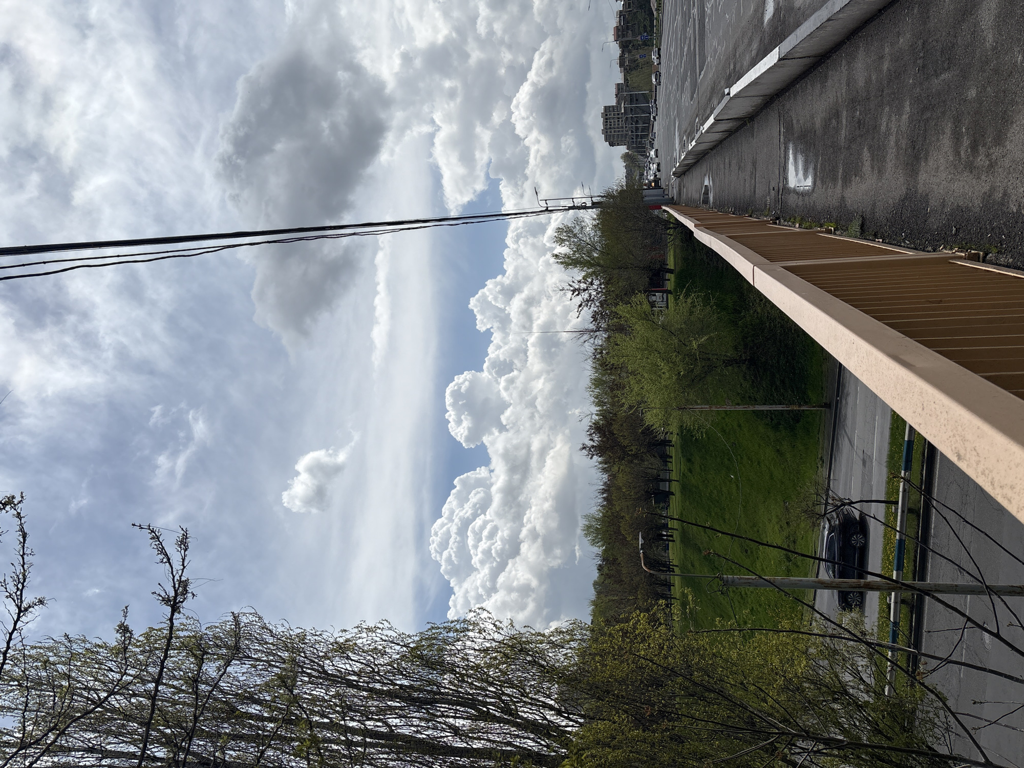 The image, taken in Yerevan, Armenia, shows a view from an elevated position, possibly a bridge or building, looking out over a landscape that includes a park, a road, and distant buildings under a cloudy sky. The foreground features a section of a metal railing and trees with budding leaves. Power lines stretch across the top left of the frame. Below, a green park area is visible, bordered by trees. A dark-colored car is parked on a road running parallel to the park. The road extends towards the horizon, where buildings can be seen in the distance. The sky is filled with cumulus clouds, suggesting a partly cloudy day.