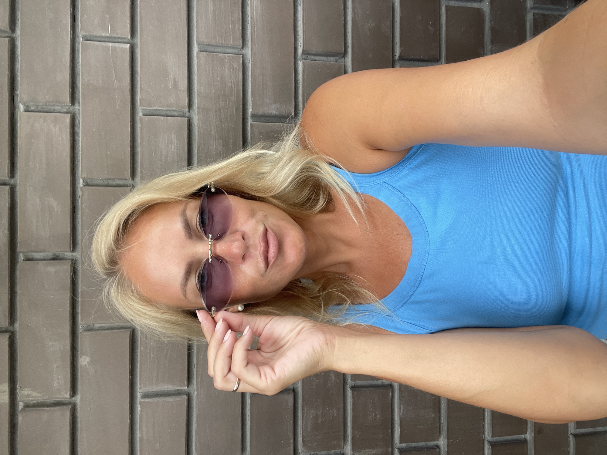 A woman with blonde hair and sunglasses is standing in front of a brick wall. She is wearing a blue tank top and a ring on her left hand. Her right hand is holding up her sunglasses. The background is a brick wall with horizontal lines. The lighting is even, and there are no shadows. The image appears to be a selfie taken outdoors. The woman is looking directly at the camera. Her lips are slightly parted, and she has a neutral expression. The overall scene is casual and informal.