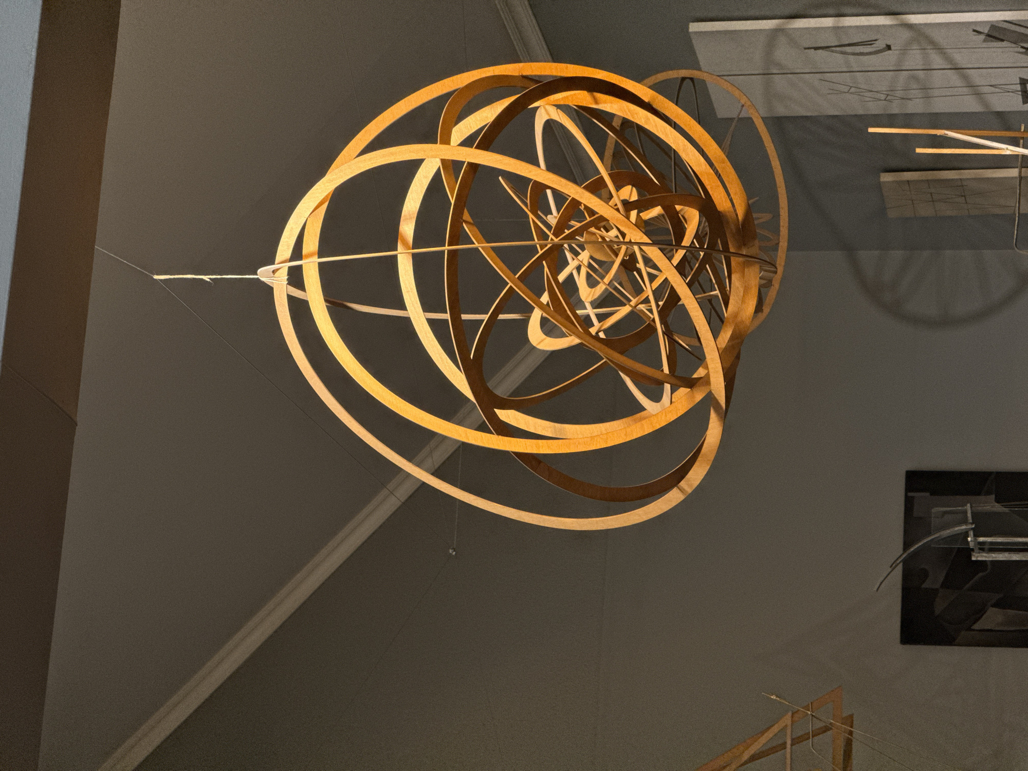 The image depicts a complex wooden sculpture suspended in an art gallery, likely indoors, given the controlled lighting and wall-mounted artwork. The sculpture consists of intersecting wooden rings, forming a spherical or elliptical configuration. It appears to be suspended by a thin wire or string. In the background are gray walls with other artworks and architectural details, including a framed piece of artwork on the right side of the frame. There is no information about the location in the city of Moscow, Russia, or people.