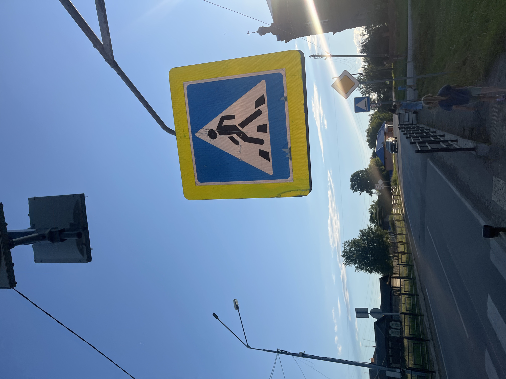 This image depicts a street scene in the village of Brusnyatskoye, Russia, under a clear blue sky with some wispy clouds. A prominent pedestrian crossing sign, featuring a silhouetted figure walking across stripes within a blue triangle bordered by yellow, is prominently displayed, hanging from an overhead structure. In the foreground, a paved road with white lane markings stretches into the distance. To the right of the road, a sidewalk is visible, with a black metal fence bordering it. Several children are present on the sidewalk, with one young girl in the foreground facing away from the camera. In the background, traditional wooden houses with dark roofs line the street. A street lamp stands tall on the left side of the road. Further down the road, another diamond-shaped warning sign and a blue square sign are visible. A dark-colored car is driving on the road. The overall environment suggests a rural or semi-rural setting in Russia during daylight hours.