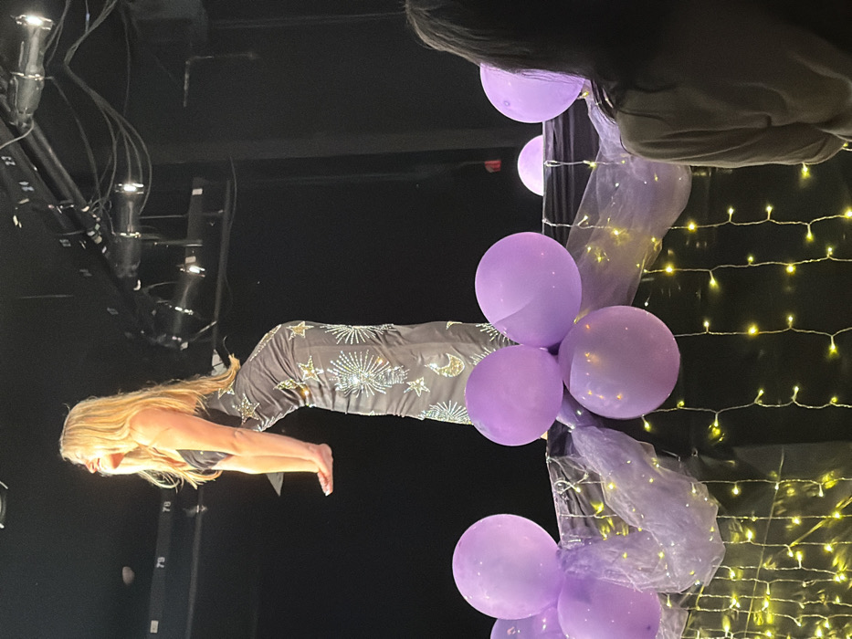 An indoor stage event is depicted in Palos Verdes Estates, United States. A blonde female performer is positioned center-stage, captured in profile from the upper torso to the knees, leaning forward with her head tilted back and a smiling expression. She wears a dark, form-fitting gown adorned with silver-colored star, sunburst, and crescent moon embellishments. Her arms are bent at the elbows with hands positioned forward. The background is uniformly dark, revealing overhead stage lighting fixtures, cables, and numbered structural supports (e.g., "24," "25," "27").
In the foreground, a decorated barrier separates the stage from the audience. This barrier is draped in dark fabric, interwoven with light purple sheer material and illuminated by multiple strings of warm white fairy lights. Several light purple balloons are clustered on top of the barrier, and two spherical, illuminated objects are also visible. The back of a person's head and shoulders, indicative of an audience member, is partially visible in the lower right foreground.