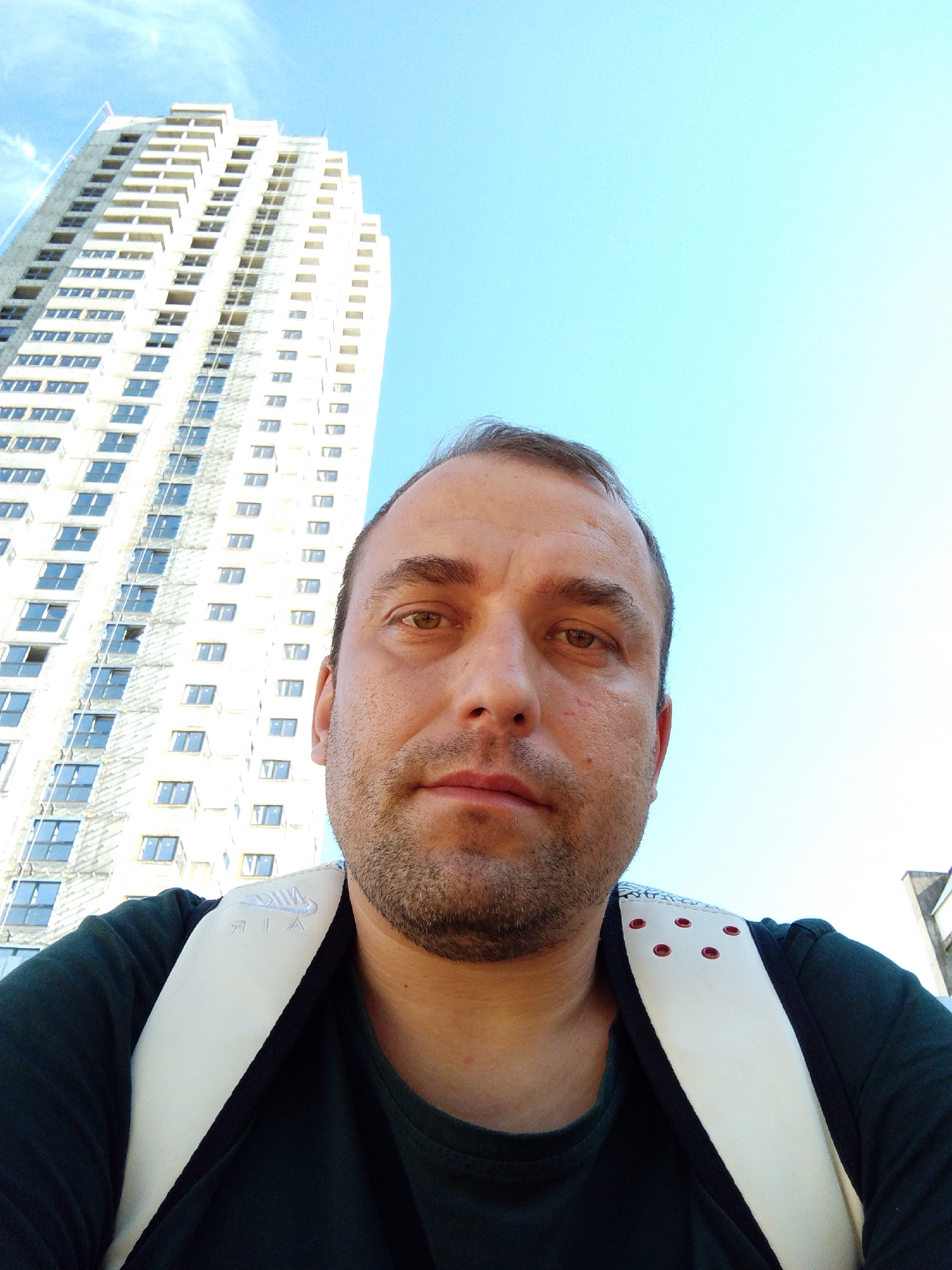 This media file is a close-up, low-angle selfie of a man with light skin and short brown hair, taken outdoors. He is looking directly at the camera with a neutral expression. He is wearing a dark green t-shirt and a white backpack with black trim. The backpack straps have a white band with the embroidered text "NIKE AIR". There are also small red decorative elements on the top of the backpack straps.
In the background, a tall, light-colored building, appearing to be under construction, dominates the left side of the frame. The building has many windows, most of which are dark, suggesting they are empty or covered. The sky is a clear, bright blue, indicating it is daytime.
The image appears to have been taken in Ufa, Russia. The sun is bright, casting minimal shadows and suggesting a clear, sunny day, likely during daylight hours. There are no other people or significant objects in the frame besides the man and the building. The overall scene suggests a casual outdoor setting, possibly a walk or commute.