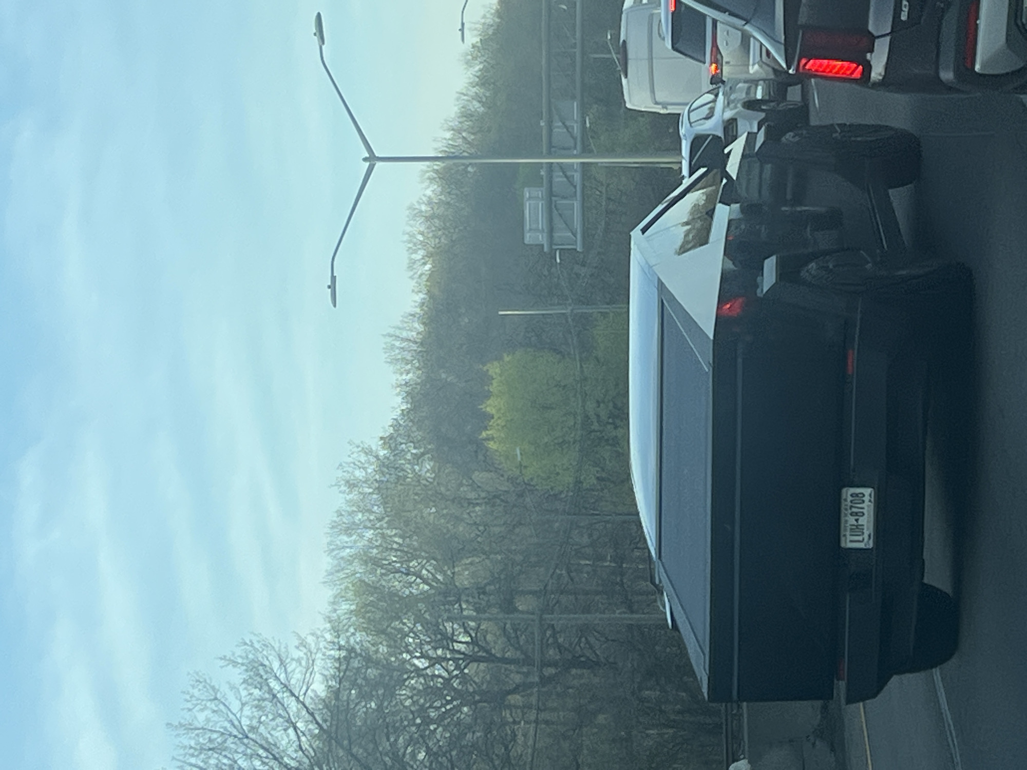 The image shows the back of a black Tesla Cybertruck on a highway in the City of Yonkers, United States of America. The vehicle has a New York license plate with the identifier "LUH-8708". The Cybertruck is among other vehicles in what appears to be a traffic jam. The background includes trees and a highway overpass structure. The lighting suggests a daytime setting with overcast conditions.