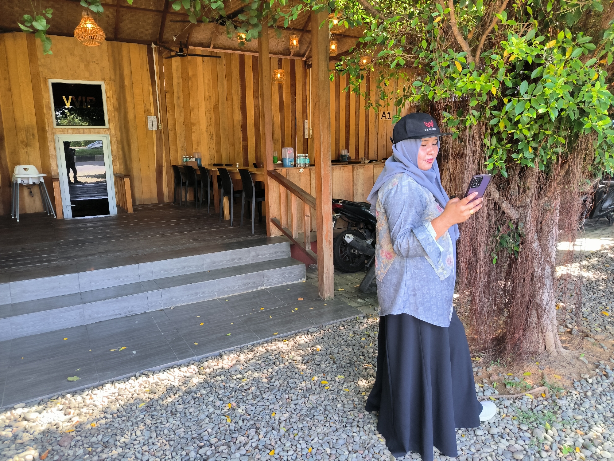 The image shows a woman standing outdoors, looking at a smartphone. She is wearing a black baseball cap, a blue hijab, a patterned blue blouse, and a long black skirt with white shoes. She is standing on a gravel surface near a tree with exposed roots.
Behind the woman, there's a wooden structure that appears to be a restaurant or cafe. The entrance is visible with a "VIP" sign on the door. Inside, there are tables and chairs. A motorbike is parked next to the wooden structure. The scene appears to be in the city of Santan, Indonesia, as indicated by the metadata.