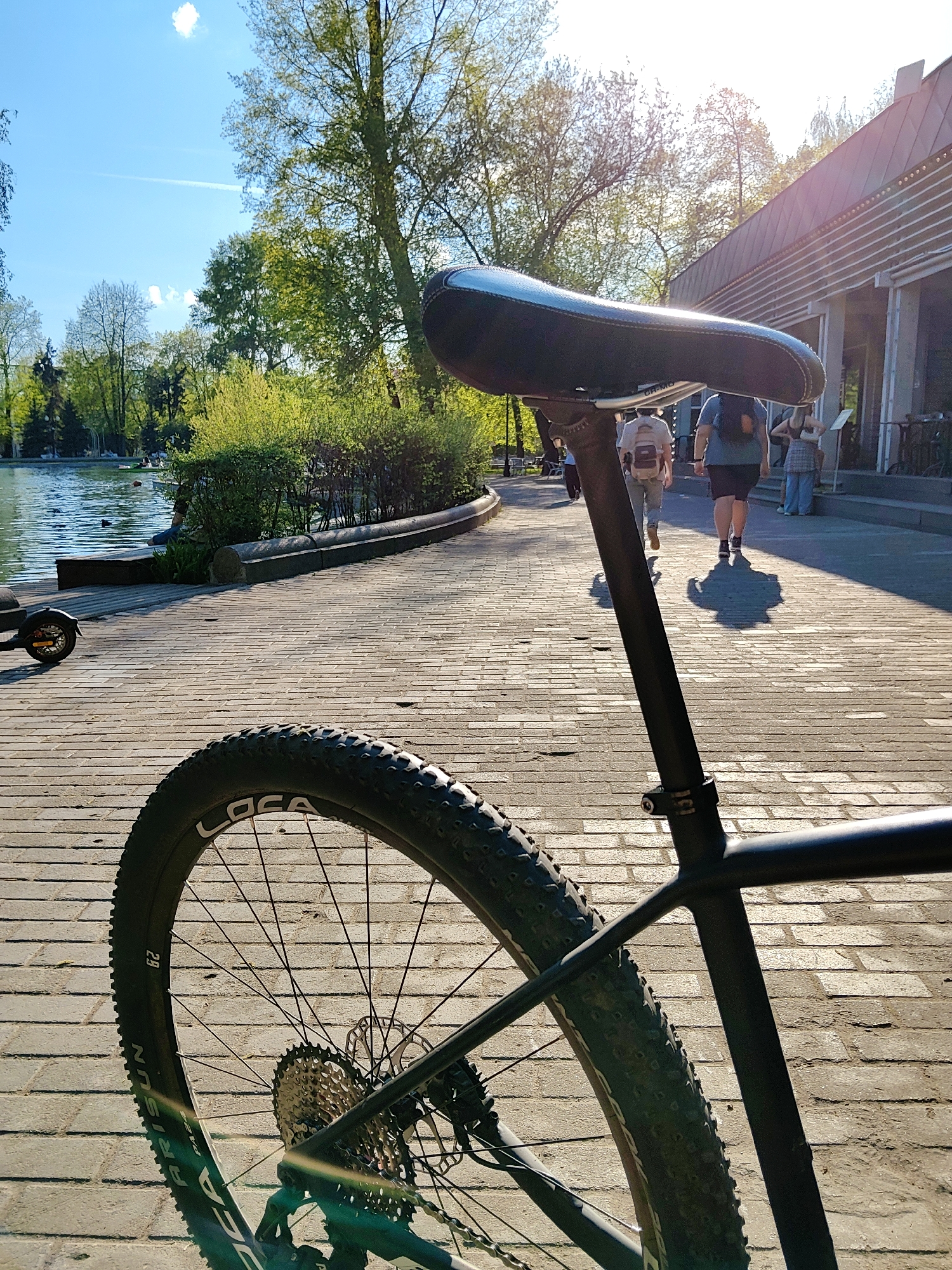 A bicycle's back wheel and seat are prominently featured in the foreground of a photo taken on a brick-paved path, likely in a park. The background reveals a lake, trees, and a building, with several people walking. The location is in Moscow, Russia.
