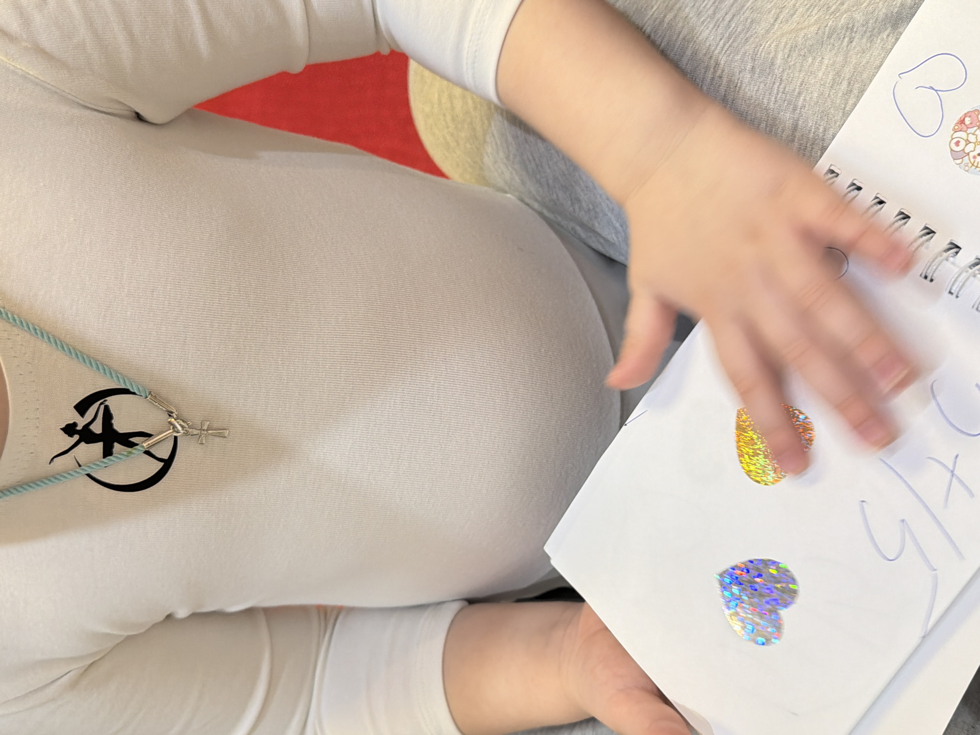The image depicts a person wearing a white long-sleeved shirt with a black logo and a turquoise necklace with a small cross. They are holding a spiral-bound notebook with a drawing and two heart-shaped stickers. The background includes a glimpse of a red surface and a gray garment. The location is not discernible from the image.