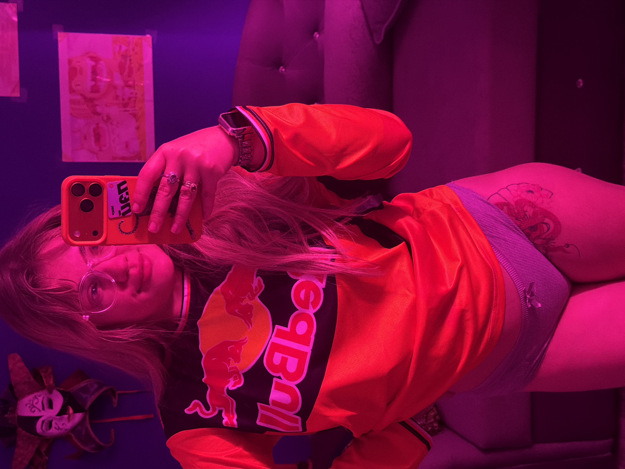 A young woman is taking a selfie in an indoor setting, seemingly in the city of Большая Шильна, Russia. She's wearing a red and black Red Bull racing jersey, eyeglasses, and several rings. She's holding an orange smartphone with a sticker on the back. Part of a mask is visible in the lower left corner. A tattoo is visible on her upper thigh. Two framed images are on the wall behind her. A tufted headboard can be seen on the right. The lighting is predominantly pink and purple.