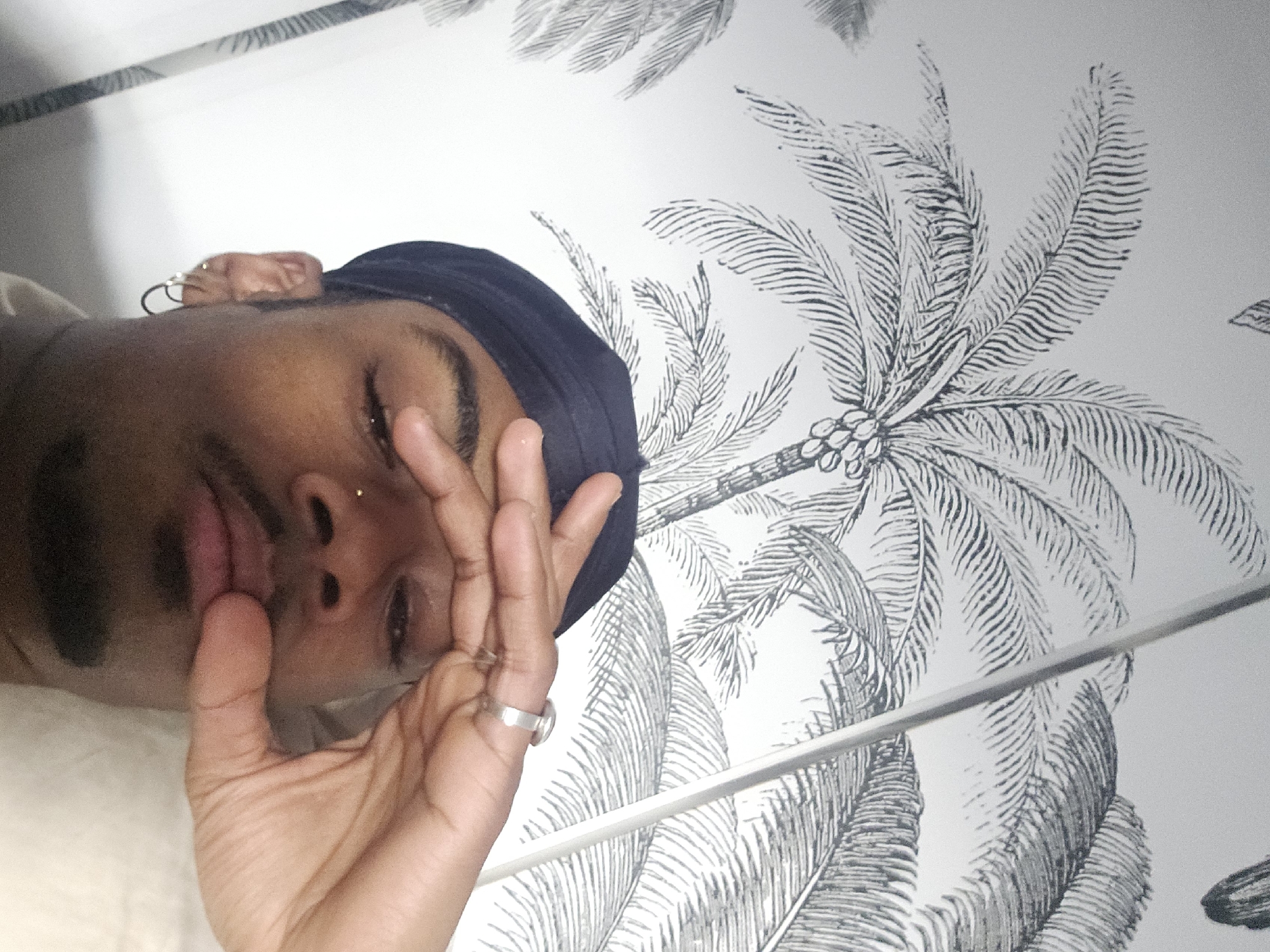 The image shows a person with brown skin and a black head covering, lying down. They have a nose piercing and are wearing hoop earrings. Their right hand is raised near their face, displaying a silver ring on the ring finger. The background features a white wall with black and white palm tree patterned wallpaper. A partial view of what appears to be a metallic rod or railing is visible in the lower right portion of the image. The location and city are unknown.