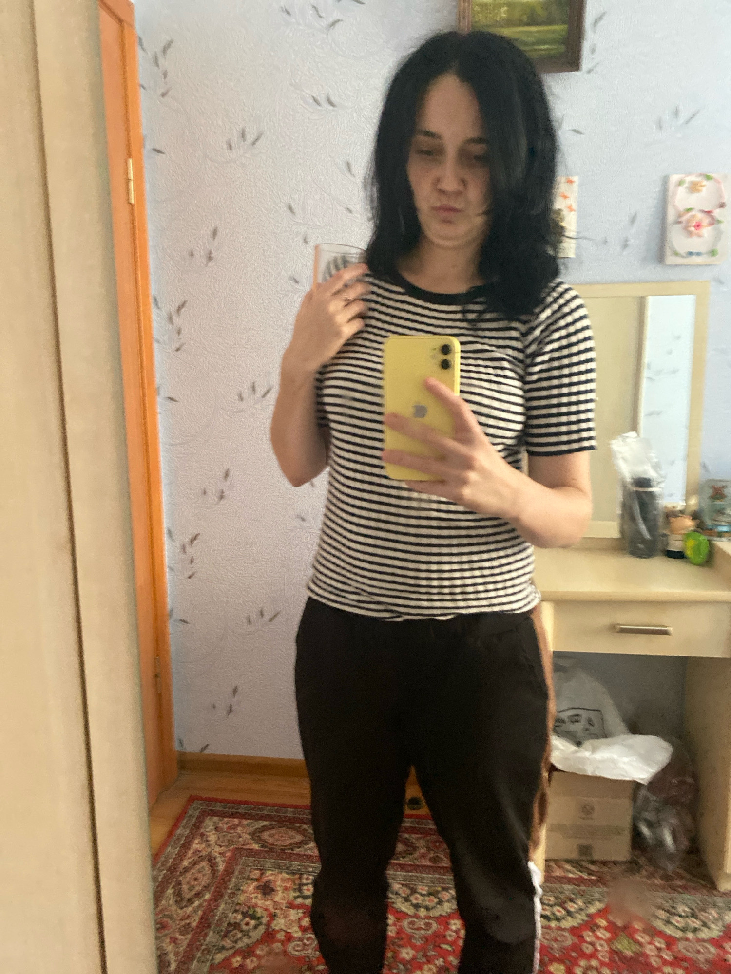 A woman is taking a mirror selfie in an indoor setting. She is wearing a black and white striped t-shirt and dark pants. She is holding a yellow smartphone in her right hand. The background includes a wall with patterned wallpaper, a wooden floor, a rug, a mirror, and a vanity table with various items on it. There is also a closet door visible to the left. The scene appears to be a bedroom or dressing area.