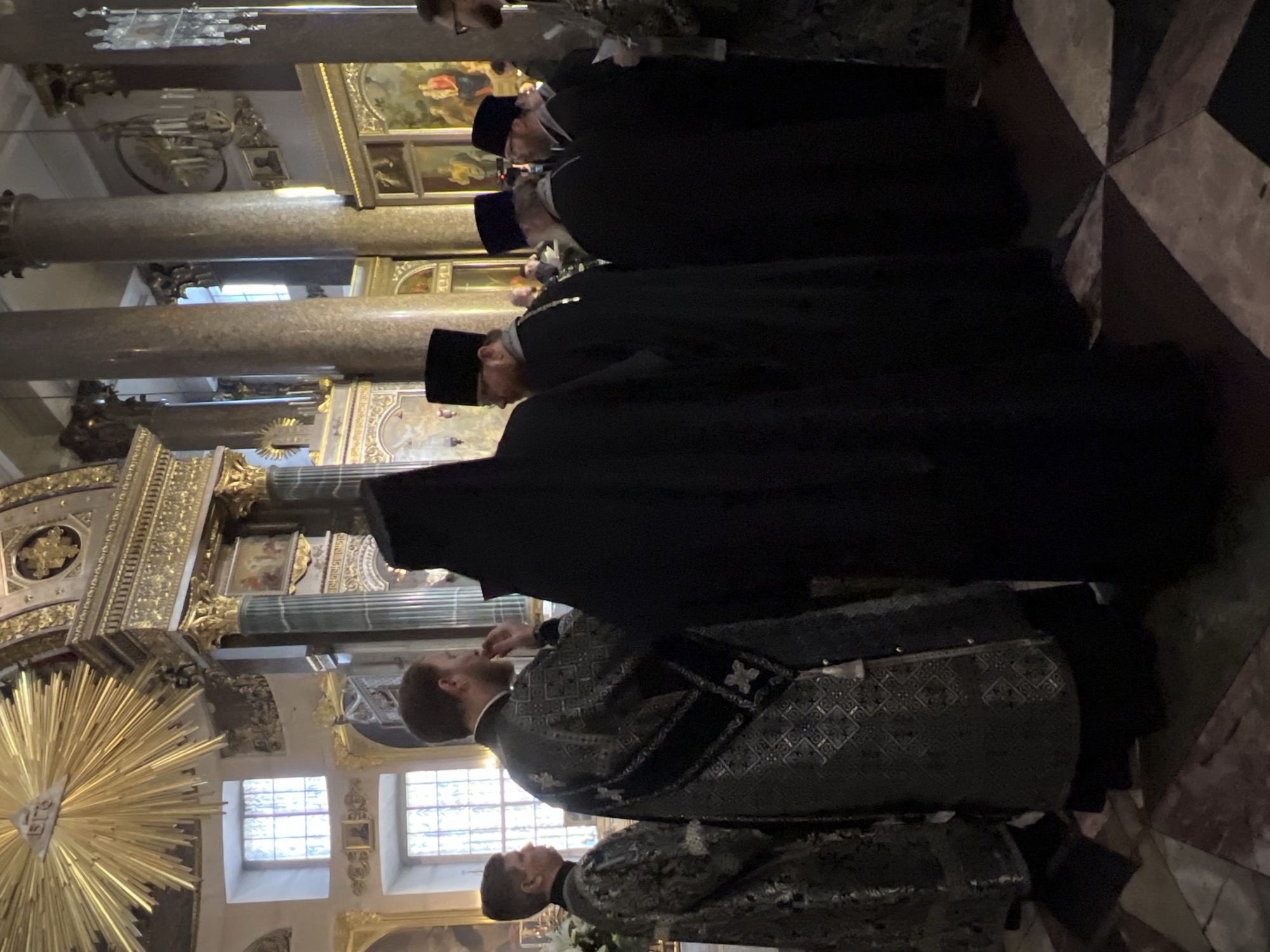 Inside the Saint Petersburg church, a group of men wearing liturgical vestments and headwear stand in a line. The group consists of a line of Orthodox priests standing in a line. The church interior includes marble columns, ornate golden decorations, and windows letting in daylight. The floor is patterned with light and dark stone tiles.