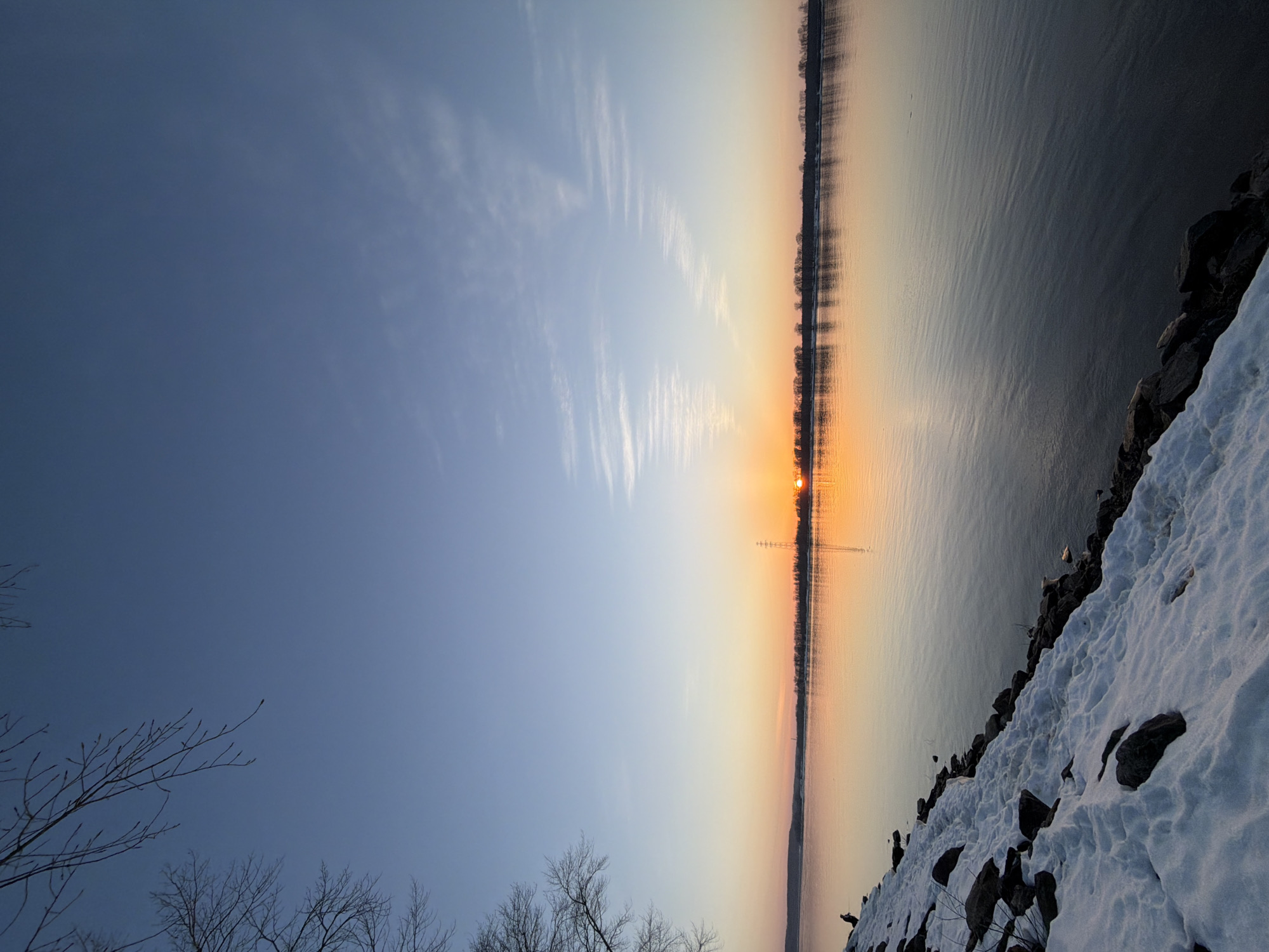 The image depicts a winter sunset over the Kama River in Chaikovsky, Russia. The sun is setting on the horizon, casting an orange glow across the water and sky. The foreground shows a snow-covered shoreline with exposed rocks. Bare tree branches are visible in the lower-left corner against the blue sky, while the horizon features a treeline on the opposite bank of the river. Power lines are visible extending over the water near the center of the image. The sky transitions from blue to orange near the horizon, with wispy clouds scattered above. The water surface reflects the sunset colors.