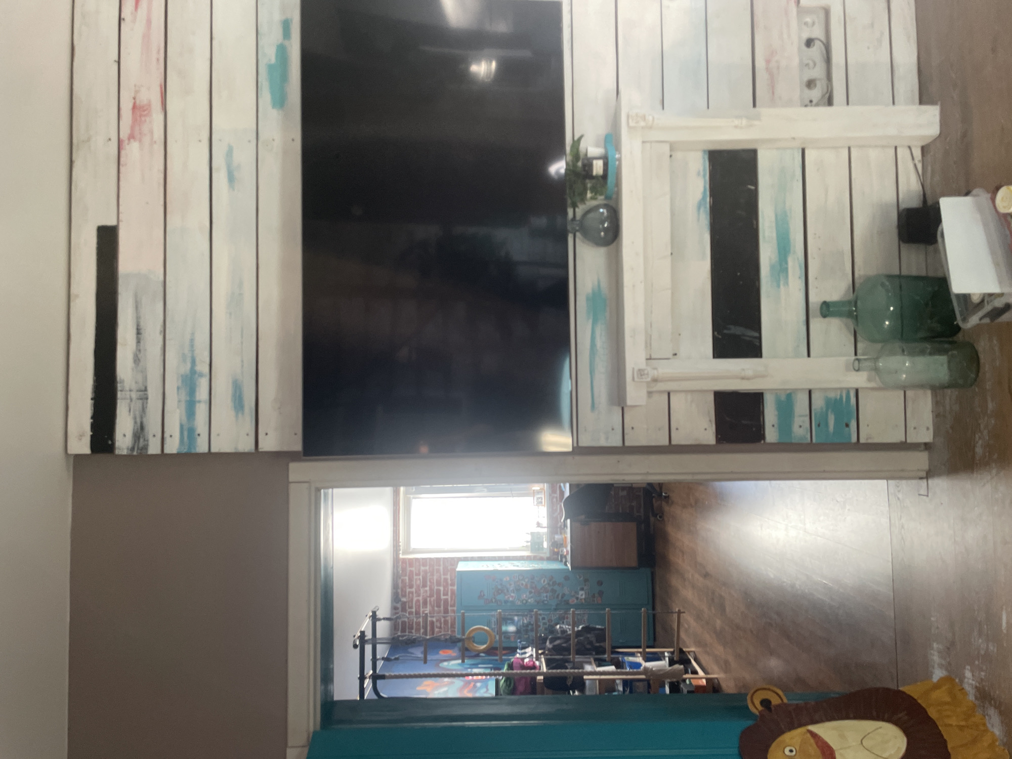 The image depicts an interior scene, likely a living room, viewed from a doorway. A wall with a rustic, horizontally paneled design, painted in white with streaks of black, red, and blue, dominates the right side of the frame. A large, dark television screen is mounted on this wall. Below the television, a white mantelpiece-like structure also features the paneled design. Two green glass bottles and a small potted plant are arranged on this mantelpiece.
To the left of the frame, a doorway opens into another room. Through this doorway, a wooden floor and a wall made of exposed brick are visible. In this adjacent room, there is a tall, teal locker, a wooden desk with a chair, and a window letting in bright sunlight. There is also a shelf unit with various items, including what appears to be a climbing rope and rings. A decorative lion cutout is partially visible on the left edge of the frame, near the doorway. The overall impression is of a lived-in, eclectic space.
The location is identified asМухавецкі сельскі Савет, Belarus.