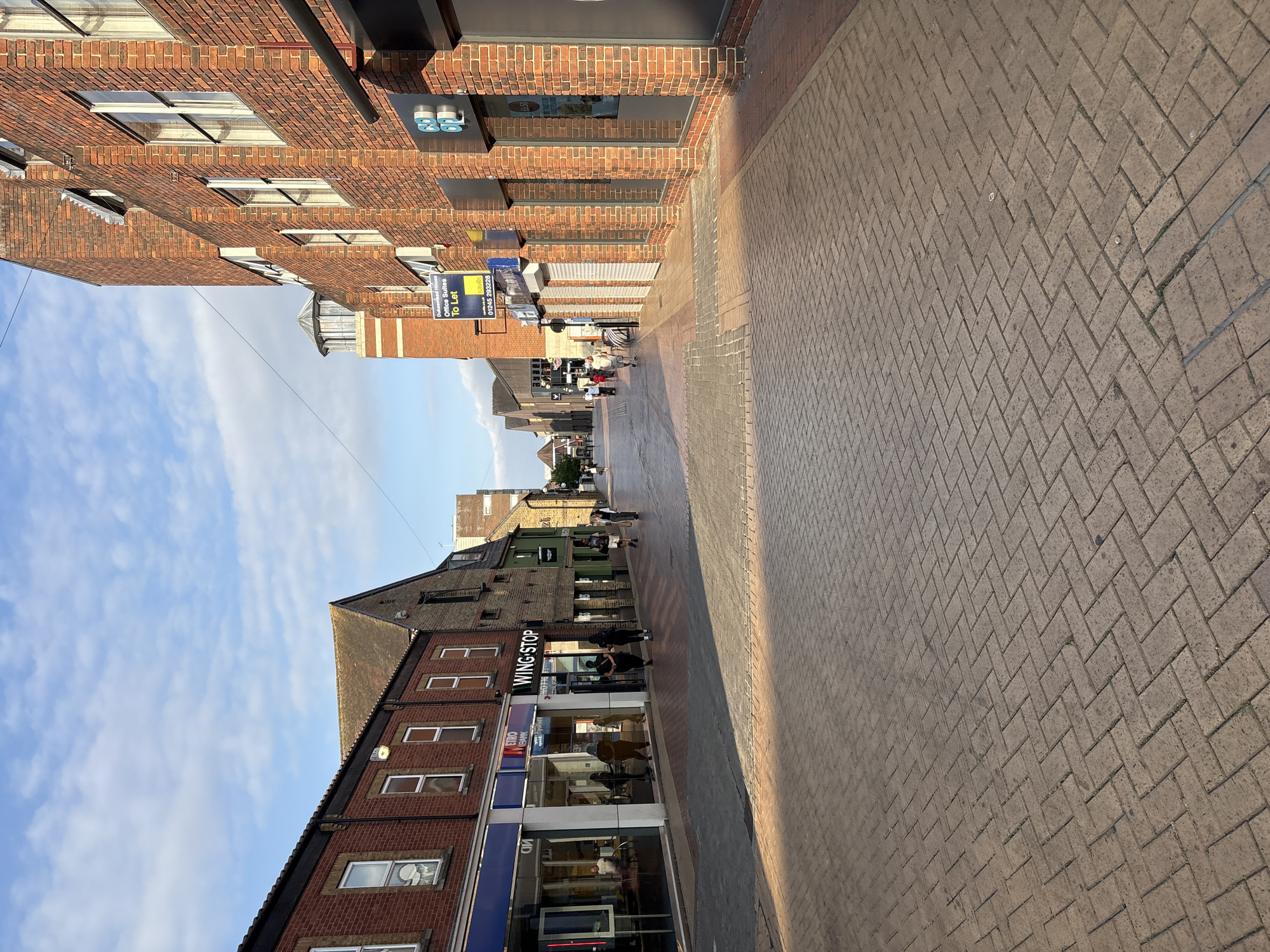 The image depicts a street scene in Chelmsford, United Kingdom. The perspective is from the street level, looking down a pedestrianized area lined with buildings. The pavement is paved with light brown bricks laid in a herringbone pattern, with patches of shadow cast by the sun. On the left side of the street, a row of shops with large glass windows is visible, including a "Wing Stop" establishment. The building on the left is constructed of red brick with white-framed windows. Across the street, on the right, is a larger brick building with multiple stories and windows. A blue sign on this building indicates "Office Suites To Let". Several people are walking along the street in various directions. The sky is blue with scattered white clouds, and the sunlight suggests it is late afternoon. The environment appears to be a commercial or retail district.