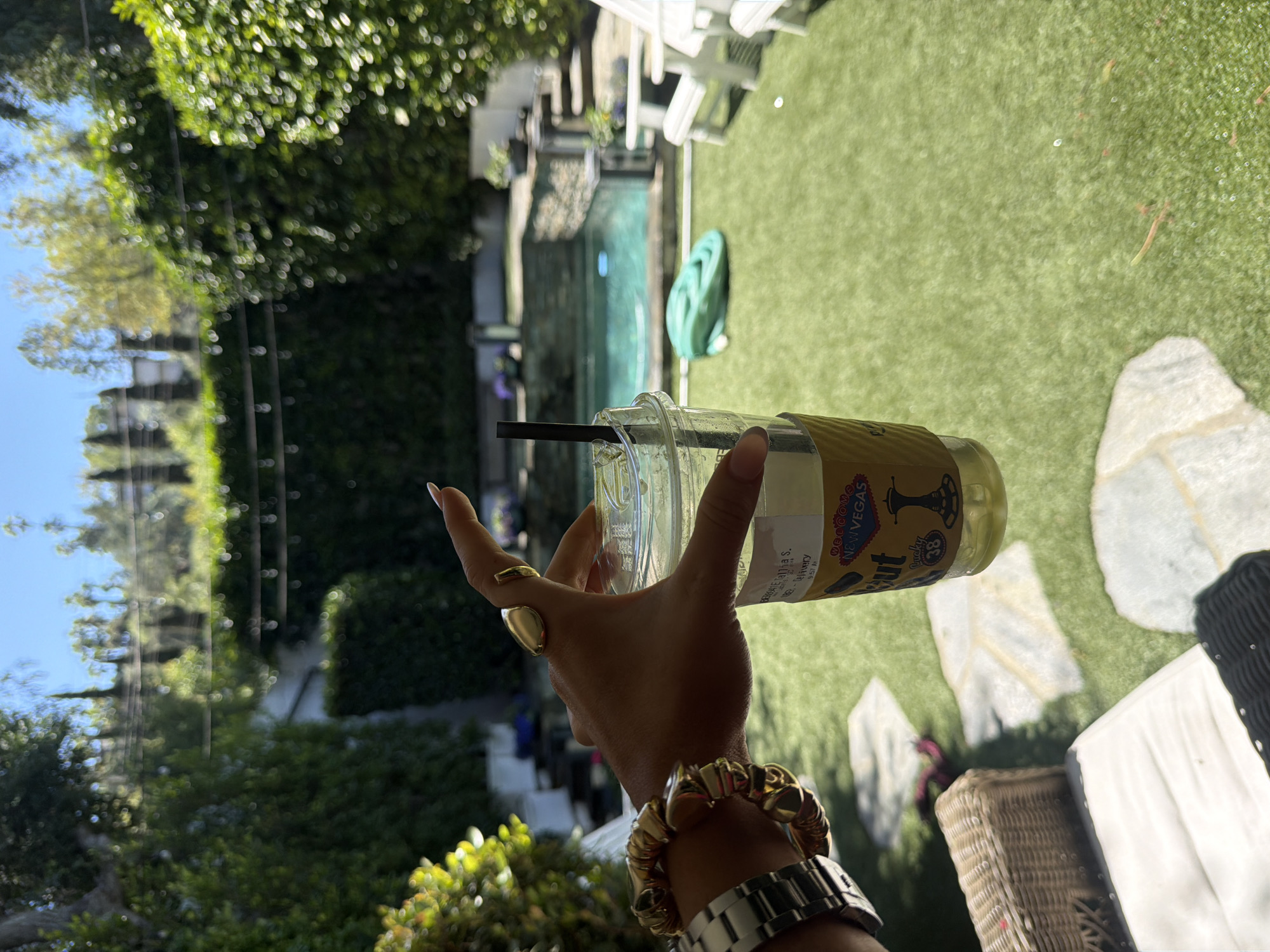 A rotated, medium-shot captures a woman's hand and forearm, adorned with two rings, a gold bracelet, and a silver wristwatch, holding a clear plastic cup with a black straw and a yellow paper sleeve labeled "WELCOME TO NEW VEGAS". The cup contains a light-colored liquid. The background is outdoors, featuring green grass, stone pavers, a partially visible wicker basket, and white outdoor furniture. A body of water, possibly a swimming pool, is visible behind the furniture. Lush green foliage covers a fence or wall, and sunlight is present, indicating daytime. The general location is suggested as Los Angeles, USA.