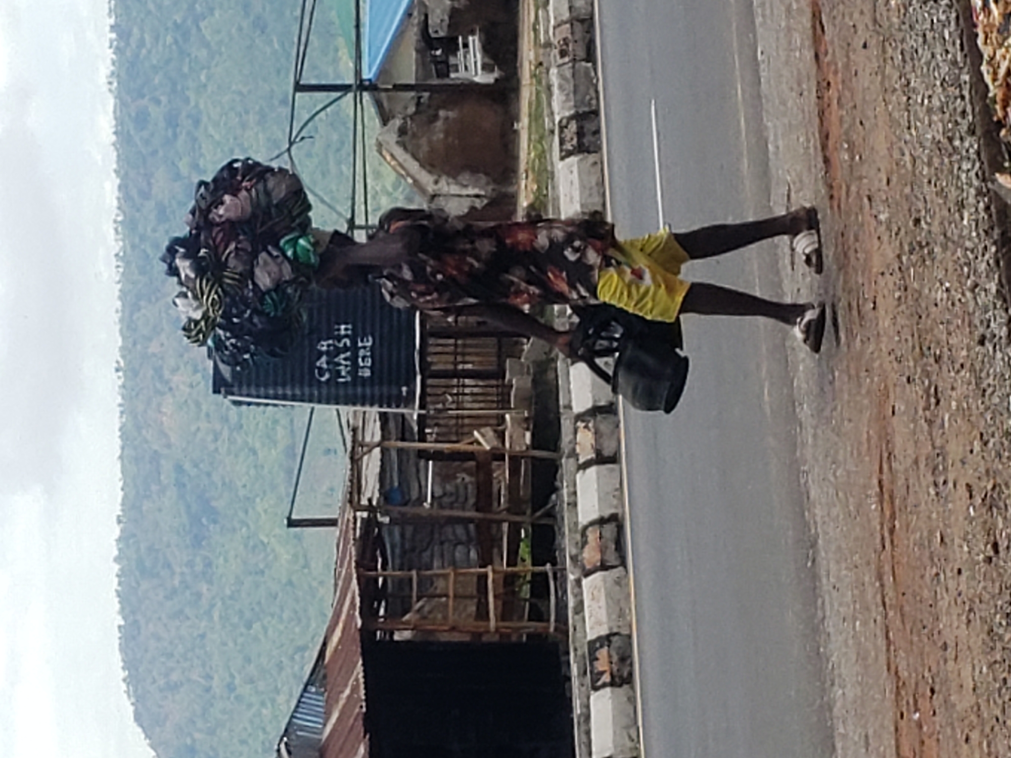 A person stands on the side of a road under natural daylight. The individual, appearing male, is wearing a patterned short-sleeved shirt, yellow shorts, and sandals. A large, bulky bundle, likely consisting of clothes or textiles, is balanced on their head. In their left hand, they hold a black bucket or bag, and a thin, light-colored pole or stick is also visible.
To the left and slightly behind the person is a tall, black plastic water tank with the words "CAR WASH HERE" clearly visible in white lettering. Further behind are structures built with corrugated iron sheets, possibly sheds or informal businesses, along with some visible wooden scaffolding.
The ground consists of a paved road directly next to the person, bordered by concrete curb stones. Beyond the curb, there's a rough gravel shoulder. In the distant background, a large, forested mountain range extends across the horizon under a cloudy sky. The scene depicts a roadside environment, suggesting daily activity or commerce in a setting consistent with Adankolo, Nigeria.