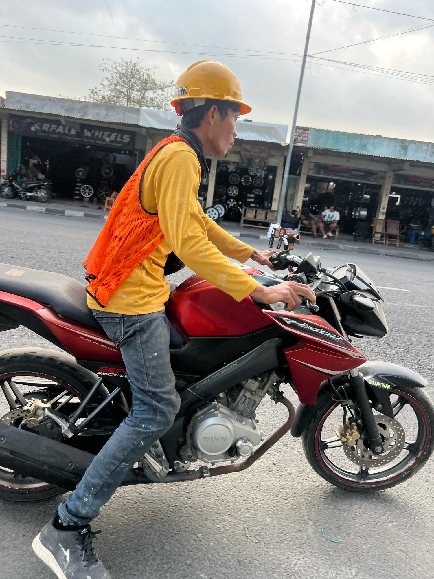 A man wearing a yellow hard hat, a yellow long-sleeved shirt, and an orange safety vest rides a red and black Yamaha motorcycle down a street in Yogyakarta, Indonesia. The man is looking to the right. The motorcycle has "VIXION" written on its side. In the background, there are several shops selling car and motorcycle wheels. The sky is cloudy.