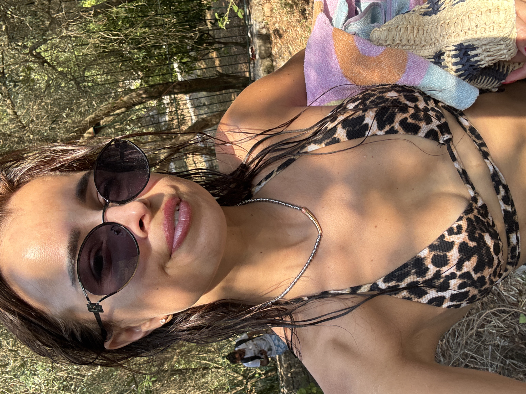 An eye-level, close-up selfie captures a woman in a leopard-print bikini and sunglasses outdoors. She is smiling and her hair is wet. She is wearing a beaded necklace and holding a colorful towel. In the background, a person with a backpack walks away from the camera. The environment appears to be a wooded area with dry foliage and sunlight filtering through the trees.