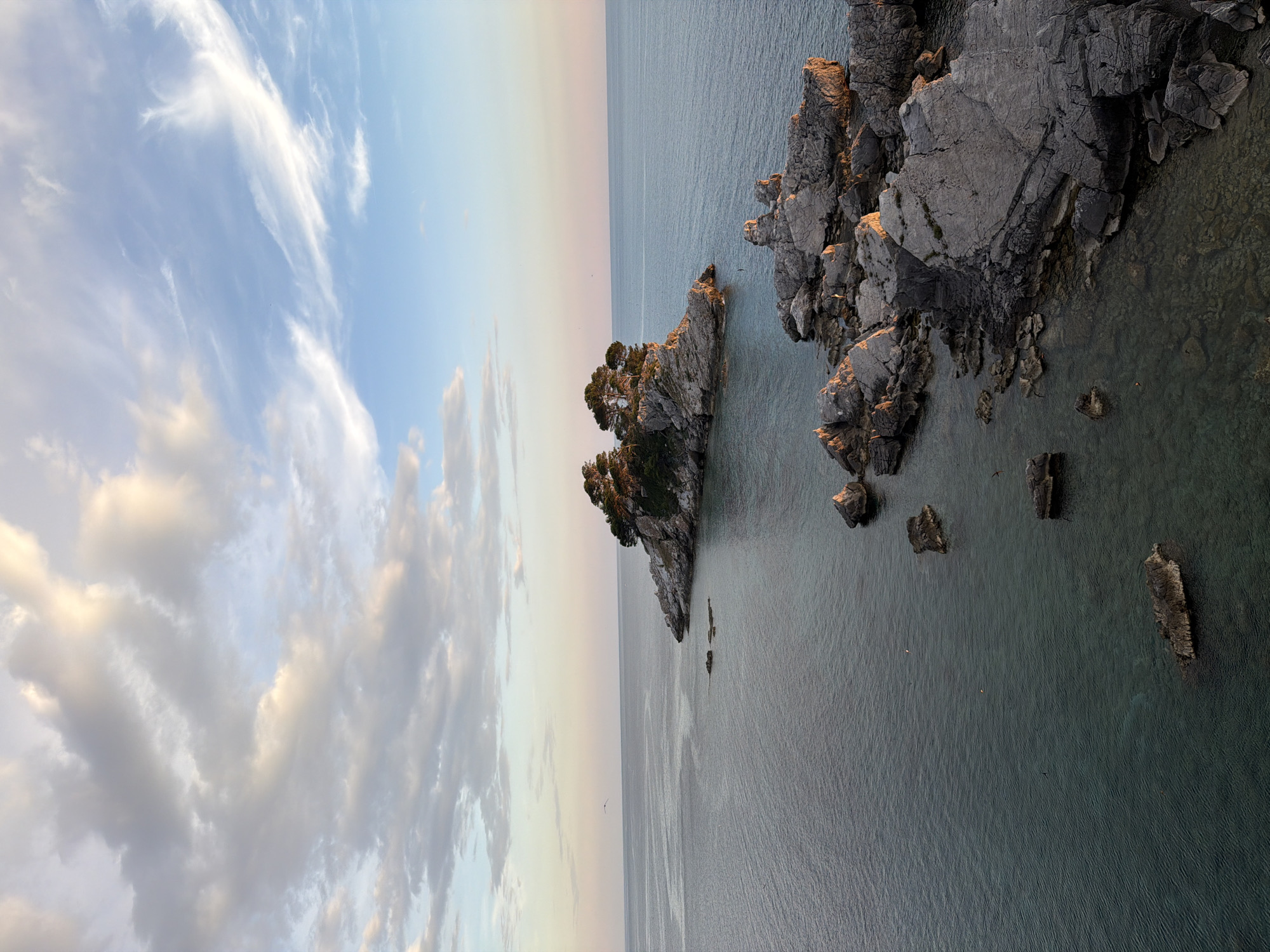 The image depicts a panoramic view of the Adriatic Sea near Petrovac na Moru, Montenegro. In the foreground, a cluster of large, weathered rocks emerges from the clear, turquoise water, with smaller submerged rocks visible beneath the surface. Beyond these rocks, a larger island formation is visible, topped with lush green pine trees. This island features a small, white-domed structure, possibly a chapel, situated on its highest point. The sea stretches to the horizon, meeting a sky filled with a mixture of soft, wispy clouds and larger, more substantial formations, painted with the gentle hues of either sunrise or sunset. The overall atmosphere is serene and natural.