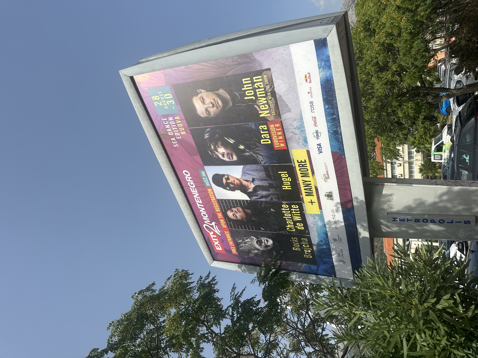 A large outdoor billboard advertising the "EXIT 2 MONTENEGRO SEA DANCE EDITION BUDBVA" festival is prominently displayed against a bright blue sky. The billboard features four artists: Boris Brejcha, Charlotte de Witte, Huge, and John Newman, with "Eurovision Winner" noted for Dara. The dates "28 AUGUST - 30" are visible. The bottom section of the billboard displays logos for sponsors including VISA, Tuborg, Coca-Cola, Rosa, Essence, and Red Bull. To the left of the billboard, a tree with green foliage partially obscures the view. In the foreground, a pedestrian signal box with the word "METROPOLIS" written vertically is visible. A street and parked cars are visible in the background, suggesting an urban setting.