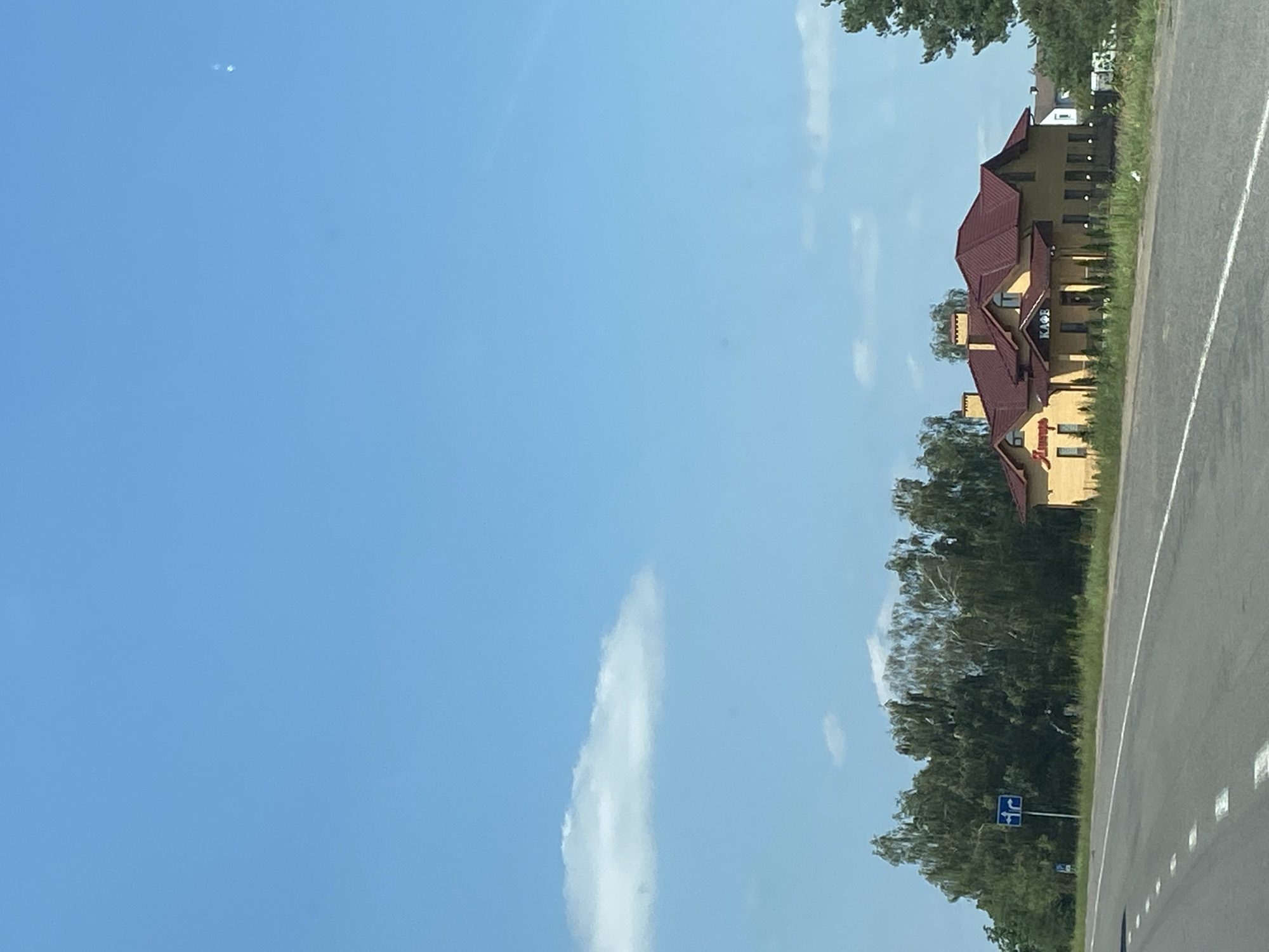 This is a photograph taken on a sunny day of a roadside building in Matykal'ski sel'ski Savet, Belarus. A paved road with white dashed lines runs across the foreground. On the right side of the road is a yellow, two-story building with a dark red roof. The building appears to be a cafe, as indicated by a sign on the front that reads "КАФЕ" (KAFE). There is also a red sign with Cyrillic lettering on the left side of the building, which appears to be a name. A line of trees, primarily birches and other deciduous trees, runs behind the building. A blue road sign with arrows indicating a turn is visible to the left of the building, in front of the trees. The sky is clear and blue with a few scattered white clouds.