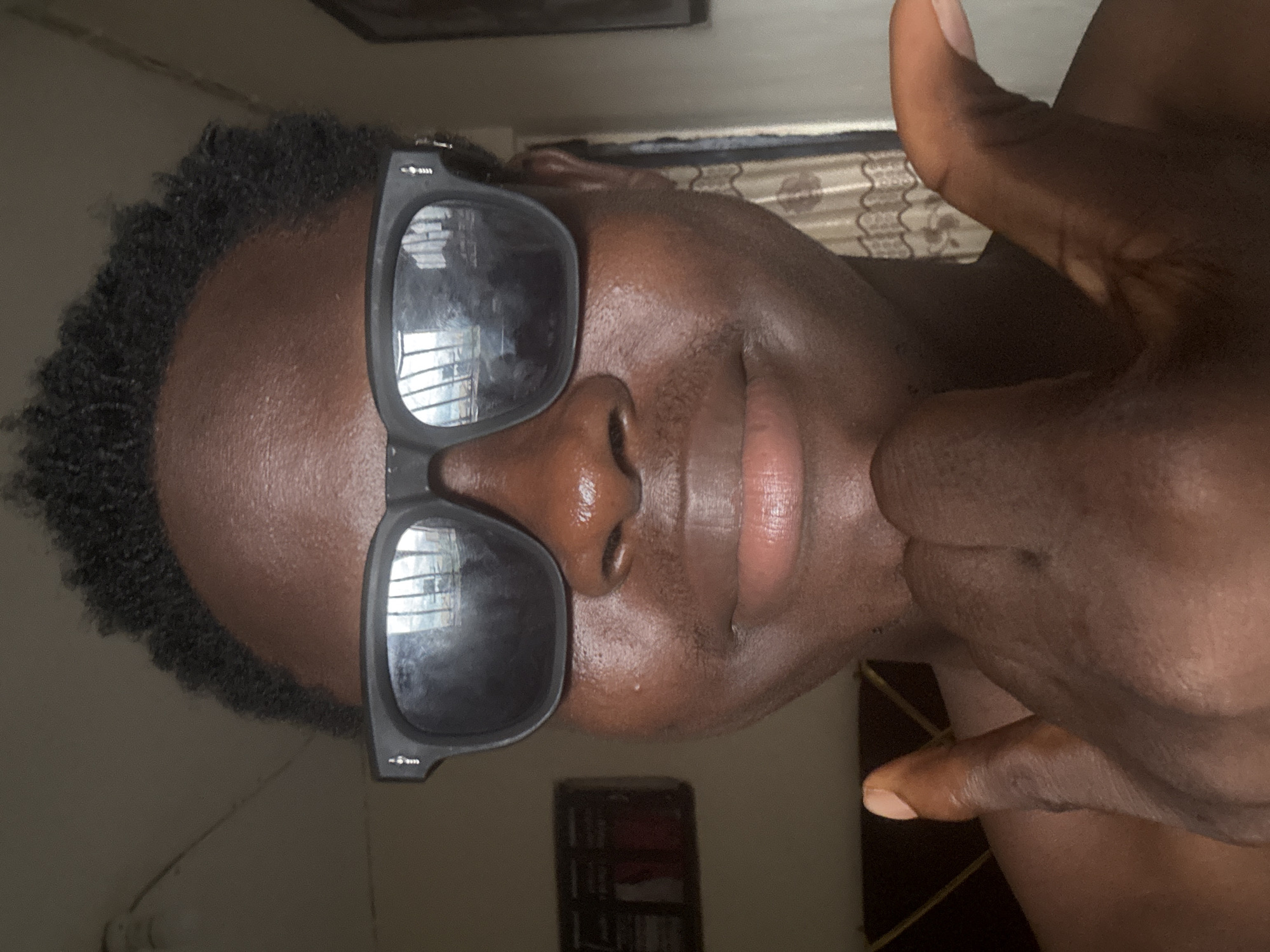 A close-up, selfie-style portrait shows a young person with dark skin wearing black sunglasses. The person's hands are visible in the foreground, with one hand resting on their chin and the other held up in a gesture, possibly a peace sign. The background is indistinct, but hints of a room are visible, including what appears to be a bed and patterned curtains. The lighting suggests the photo was taken indoors, likely during the daytime. The image is sharp and focused on the person's face and hands.
