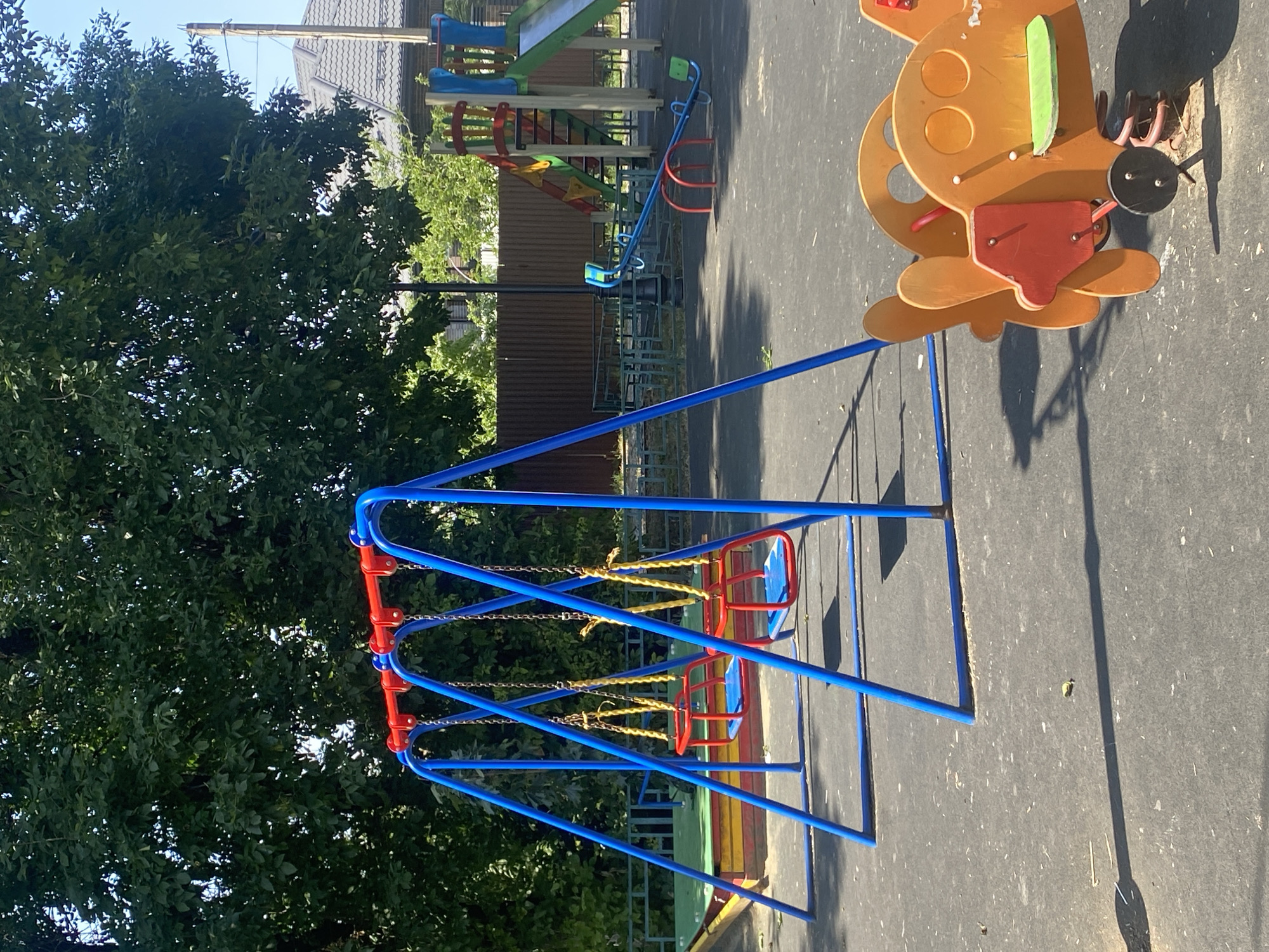 A public playground in a residential area of Zapadny, Russia is depicted. The playground features a blue swing set with red seats, a seesaw, and a brightly colored airplane-shaped spring rider. In the background, there is a large slide, a sandbox, and a wooden fence. Lush green trees and a clear blue sky form the backdrop, suggesting fair weather. The ground is paved with dark asphalt.