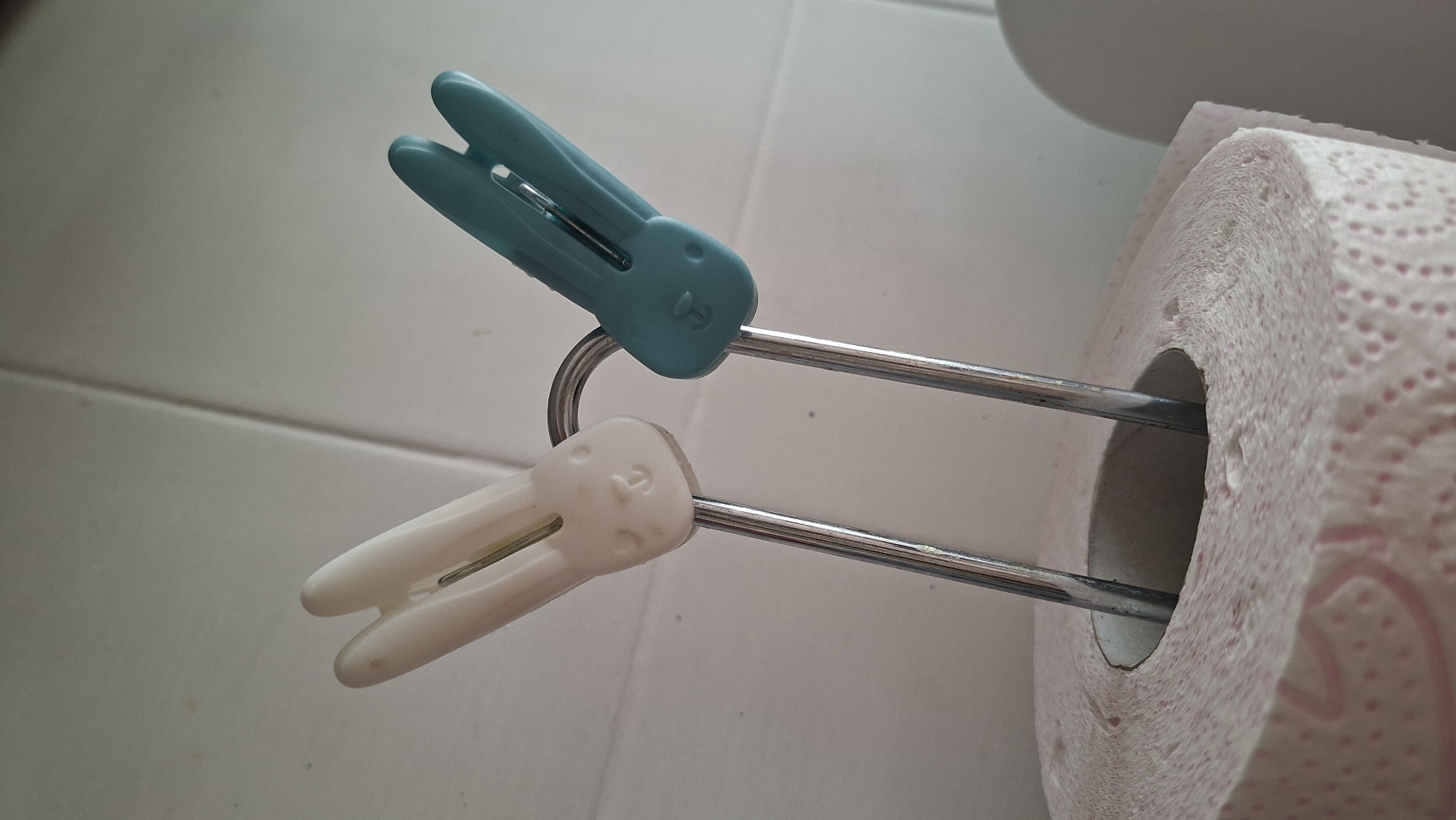 A toilet paper roll is standing upright, supported by two metal rods that are bent into a hook shape at the top. Two clothespins, designed to resemble bunny faces, are attached to the metal rods. One clothespin is white, and the other is teal. The background consists of a tiled wall. The scene appears to be in a bathroom, possibly in the city of Myadzyel, Belarus.