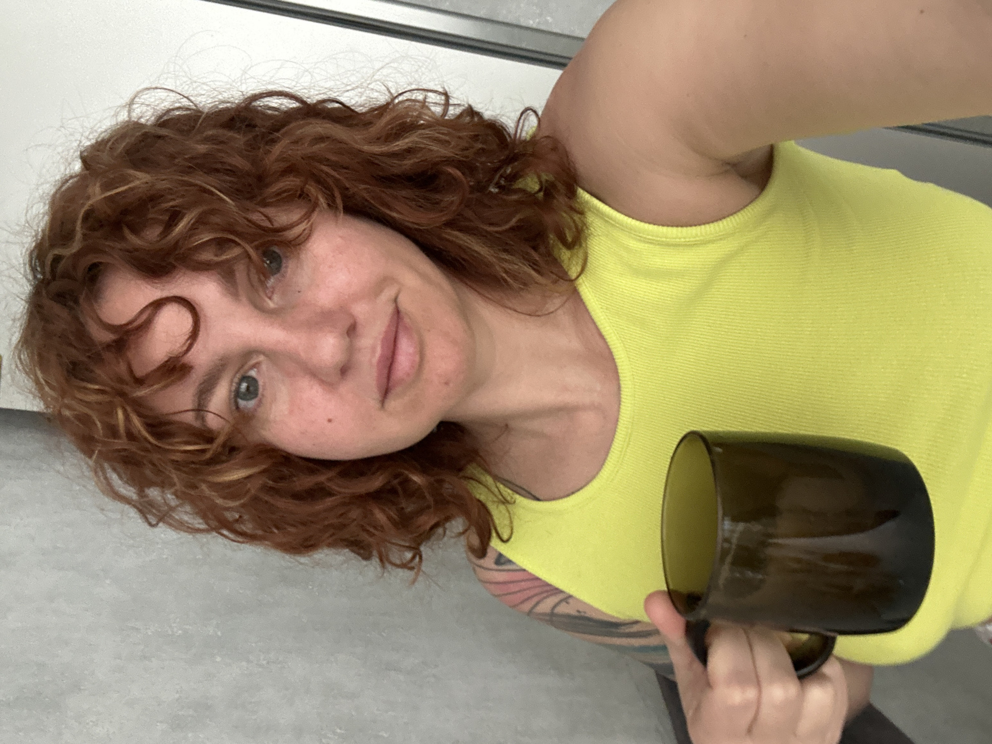 A fair-skinned woman with curly, reddish-brown hair and green eyes is holding a dark glass mug in her right hand. She is wearing a bright yellow tank top. Part of a tattoo is visible on her left arm. The background consists of a white wall and a gray floor. There is no indication of Sochi, Russia in the image.