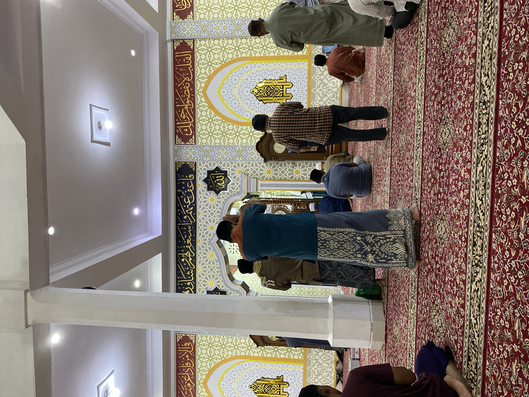 The image depicts the interior of a mosque, likely in Lhokseumawe, Indonesia, with several individuals engaged in prayer. The flooring is covered with a richly patterned red carpet. The walls are adorned with intricate yellow and white geometric designs and Arabic calligraphy. Dominating the scene are several columns and arches, characteristic of mosque architecture. In the foreground, a person wearing a dark maroon shirt and blue pants is seated. Towards the center, a person in a teal shirt and a patterned sarong stands facing away from the camera, with hands raised in prayer. Other individuals are visible in prayer positions throughout the mosque. The lighting suggests an indoor environment, possibly during a prayer service. The overall atmosphere is one of religious devotion and communal worship.